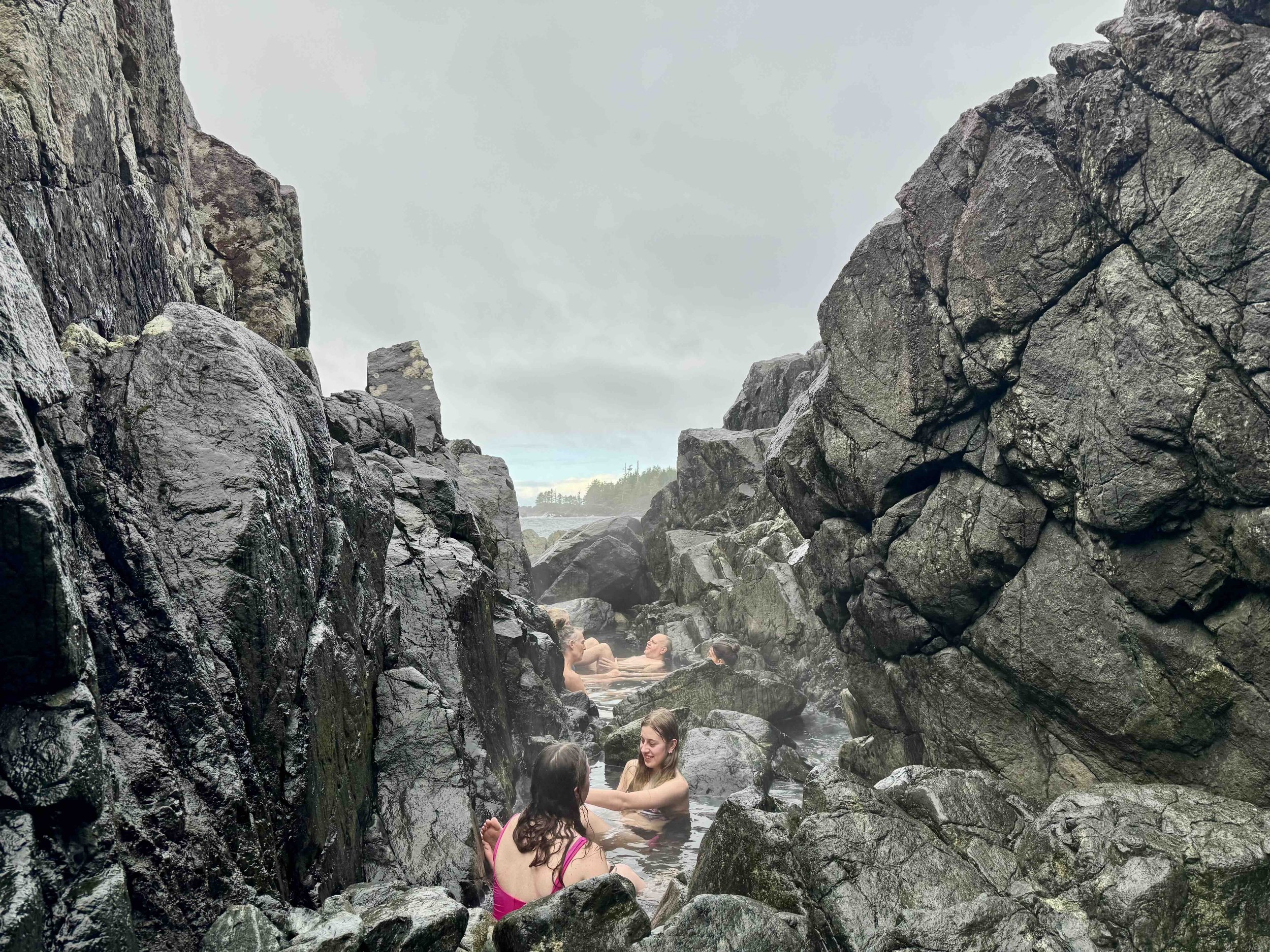 Group of people enjoying a natural hot spring between large dark rocks and cliffs, with cloudy sky and coastline in the background.