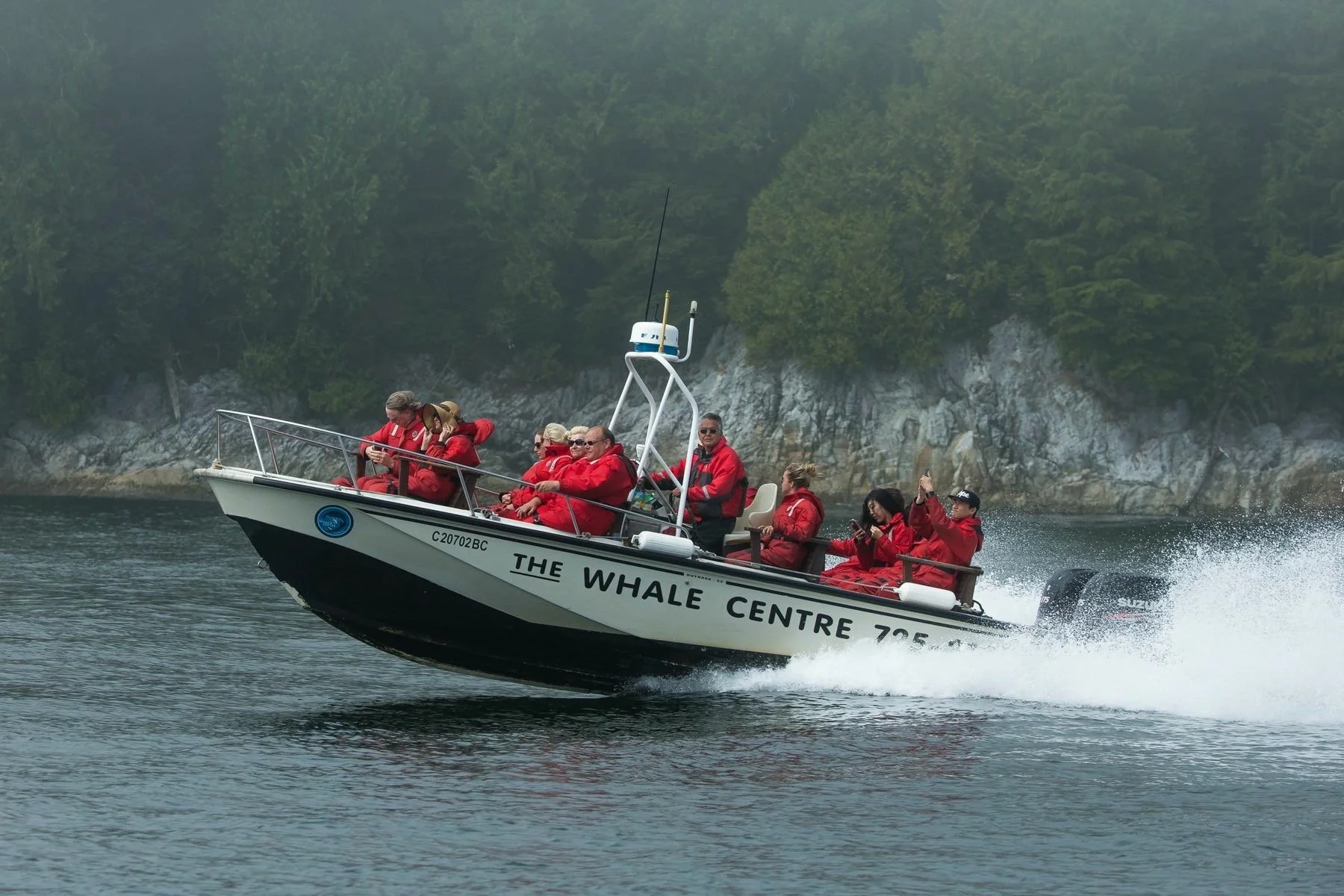 A group of people wearing red jackets on a boat speeding across water, with a forested rocky shoreline in the background.