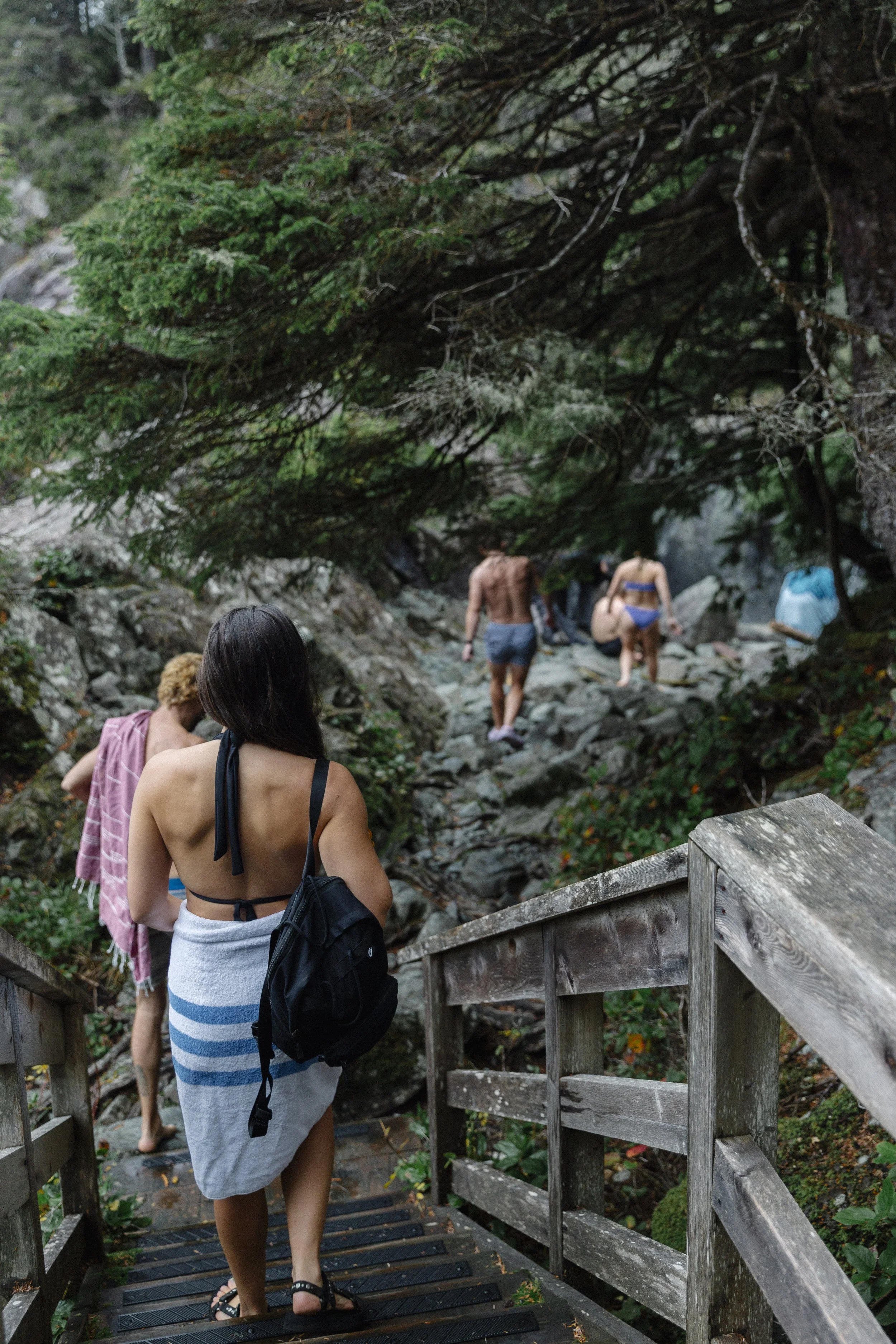 People in swimsuits hiking down rocks on a forest trail, with one woman in a towel and backpack leading.