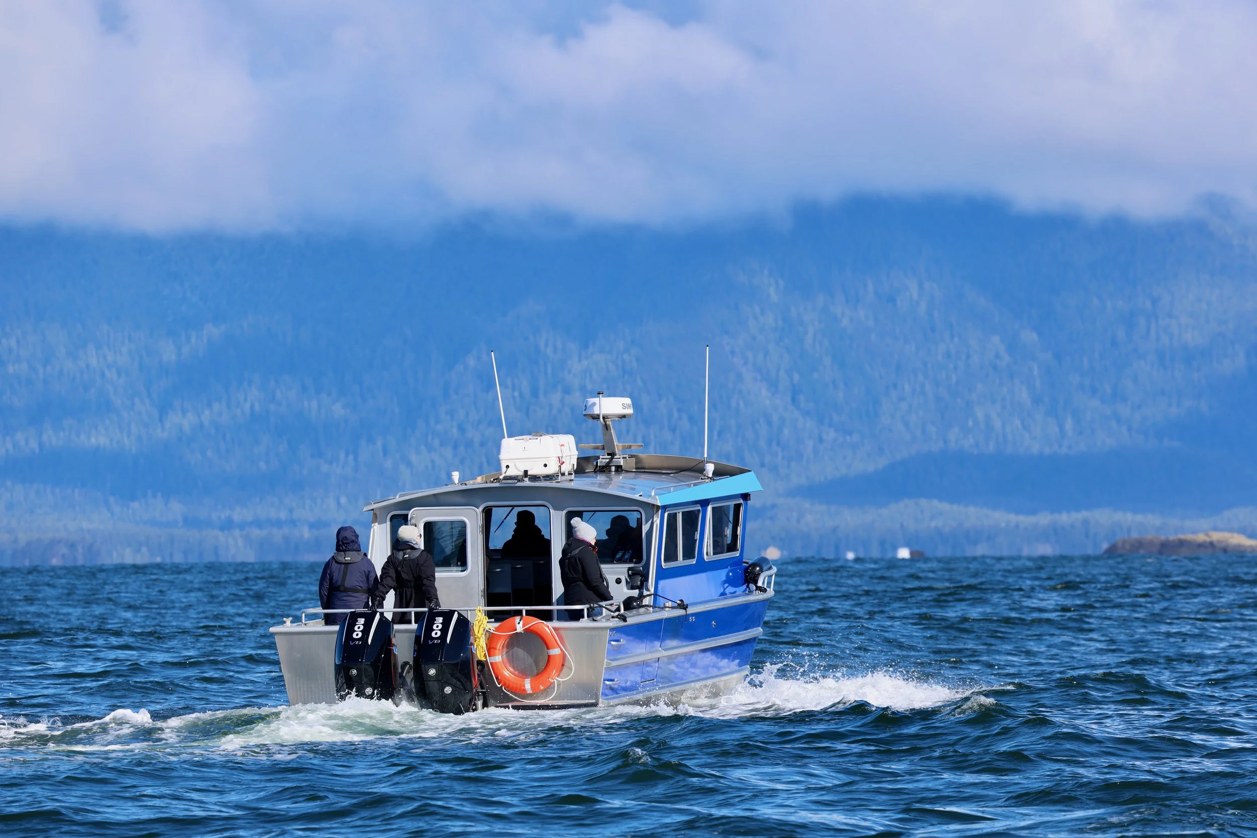 A blue and gray motorboat with four people on board, moving on the ocean with a mountain and cloudy sky in the background.