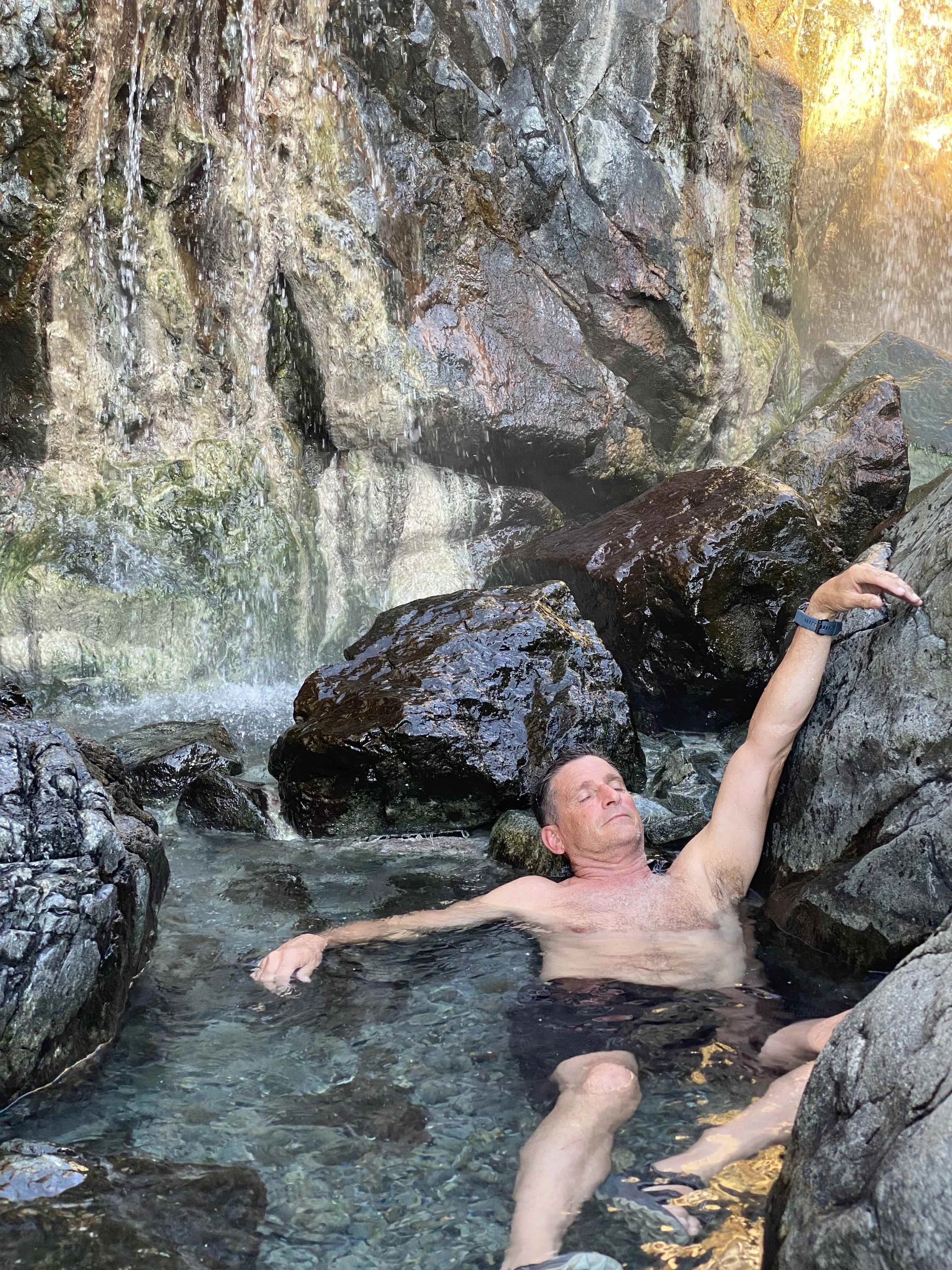 A man relaxing in a natural hot spring or creek surrounded by rocks and waterfall with sunlight shining in.