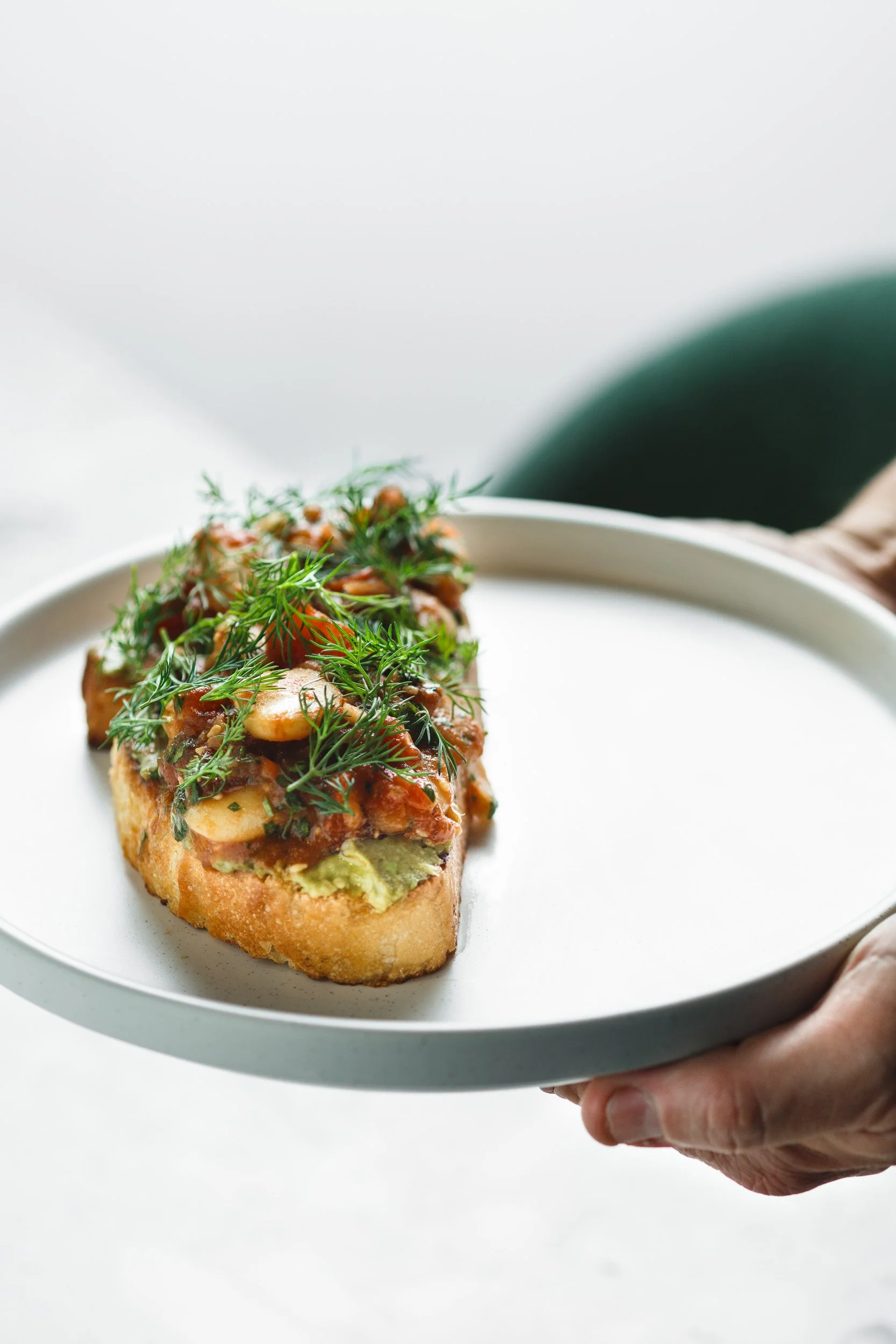  Photograph by Amy Carson, San Diego food photographer. Commercial food photo commissioned by a restaurant and hospitality group. Minimalist shot of toast with beans and avocado with fresh dill on a clean white plate held by chef’s hands. 
