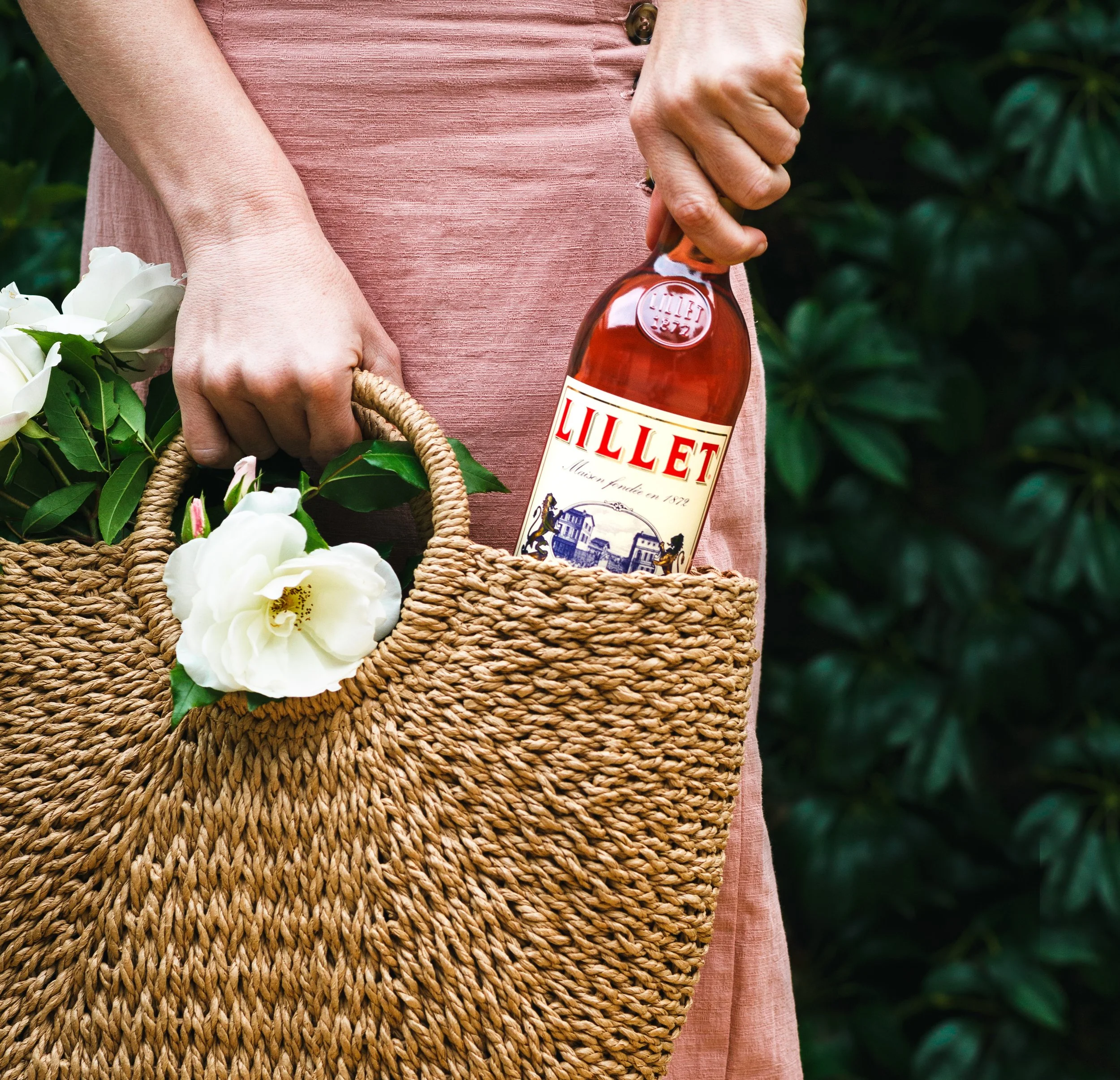  Photographed by San Diego food and drink photographer Amy Carson. Commercial lifestyle advertisement of a woman holding a woven bag with bottle of Lillet rose wine and flowers in a lush garden setting. 