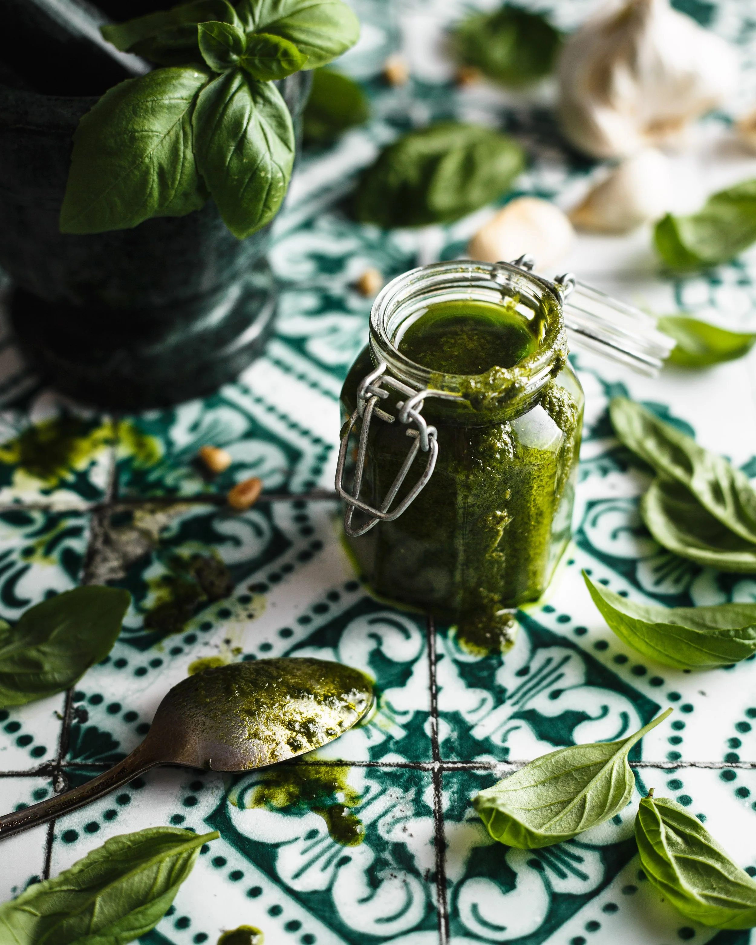  Image by Amy Carson, a food photographer in San Diego, California. Commerical food photo of pesto with fresh basil leaves and garlic with a mortar and pestle on a green tile background.  