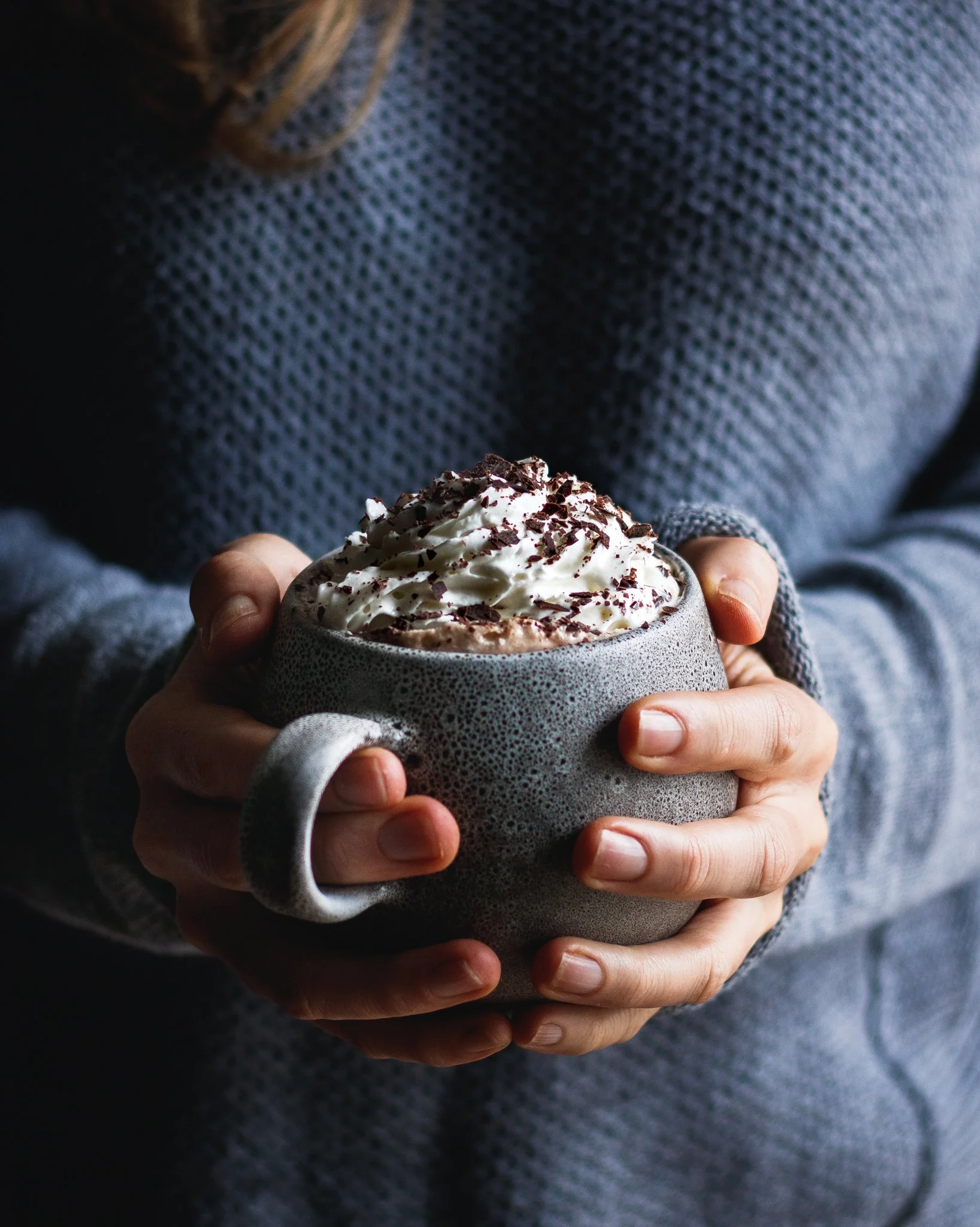  Image by Amy Carson, a commercial food photographer based San Diego. Moody editorial lifestyle photograph of woman in a gray sweater holding a mug of hot chocolate with whipped cream and chocolate shavings. 
