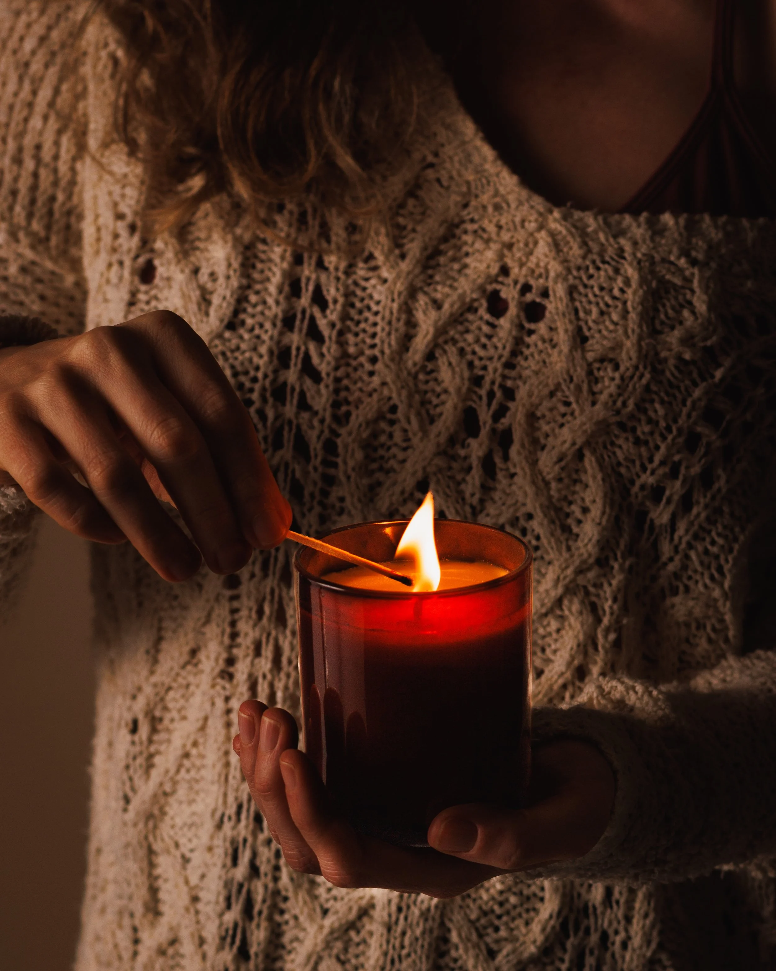 Commercial lifestyle product photo of a girl lighting a candle in a dark, cozy aesthetic.