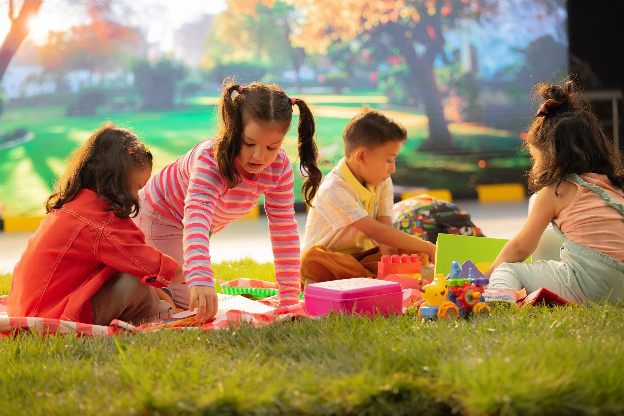 Ahlan Simsim unit still: four children play with toys and drawing books on a picnic blanket on grass.