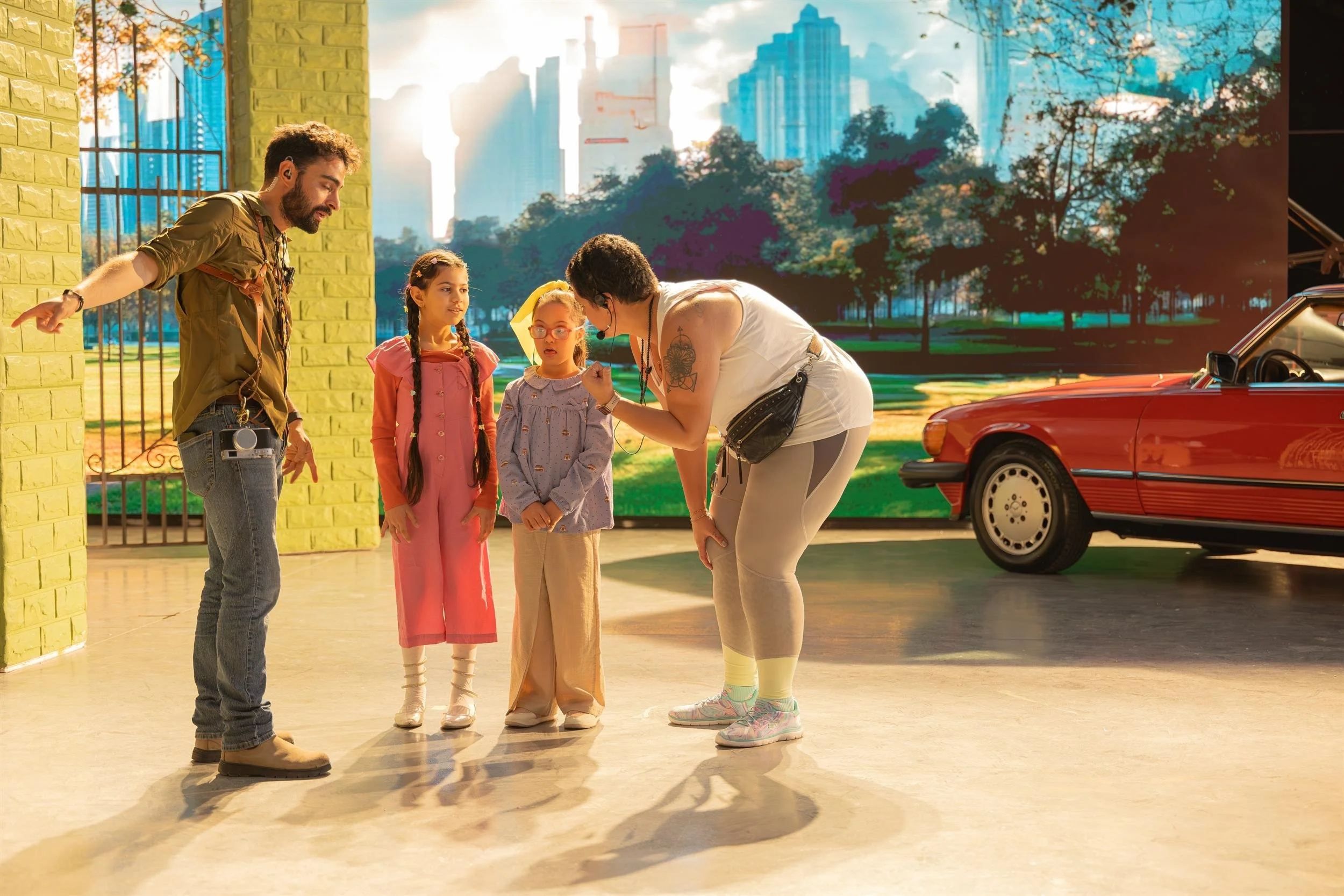 Ahlan Simsim unit still: Crew members talk with two children on a set with a city-park screen and a red car.