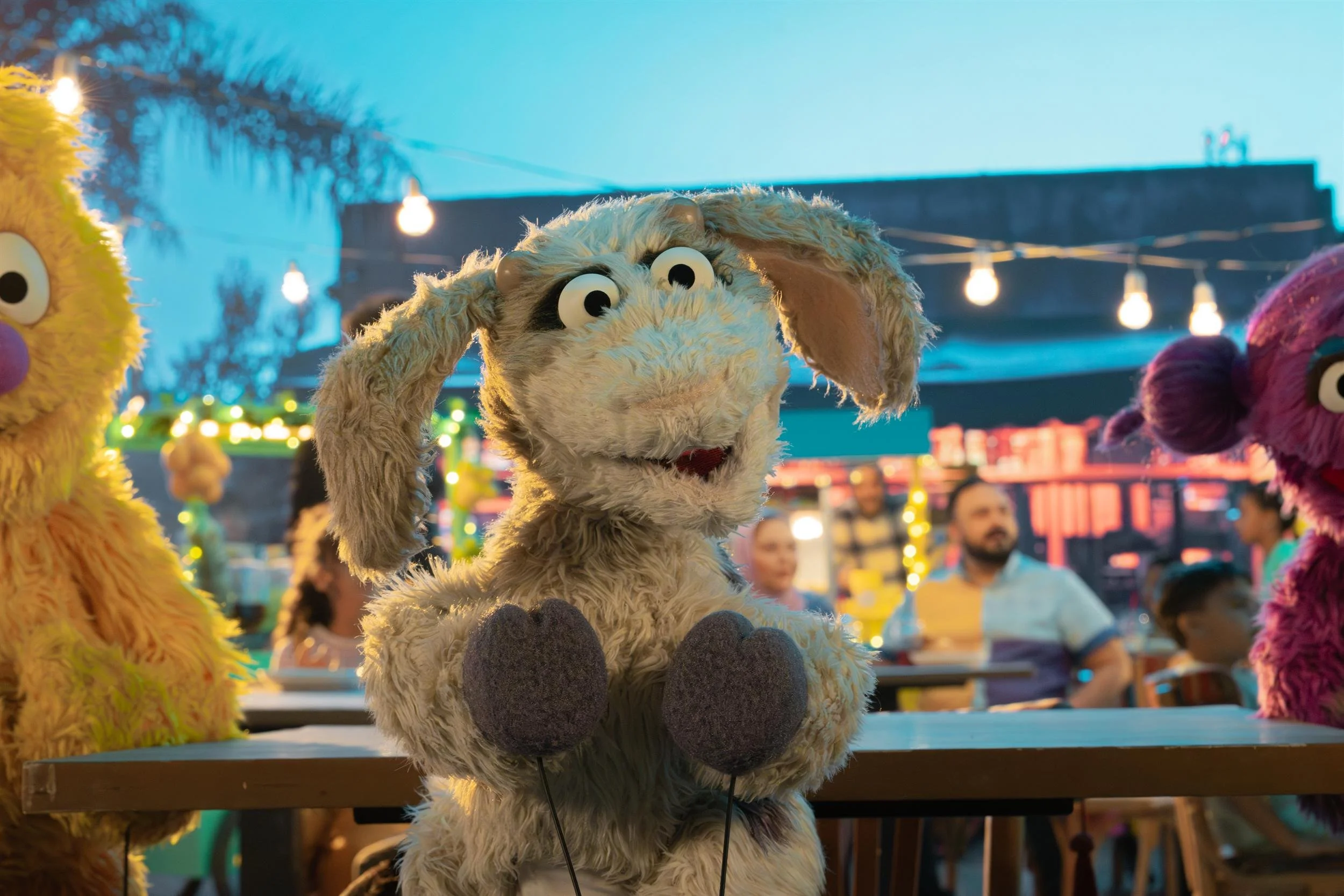 Ahlan Simsim unit still: a grey goat Muppet stands between two other Muppets at a night market.