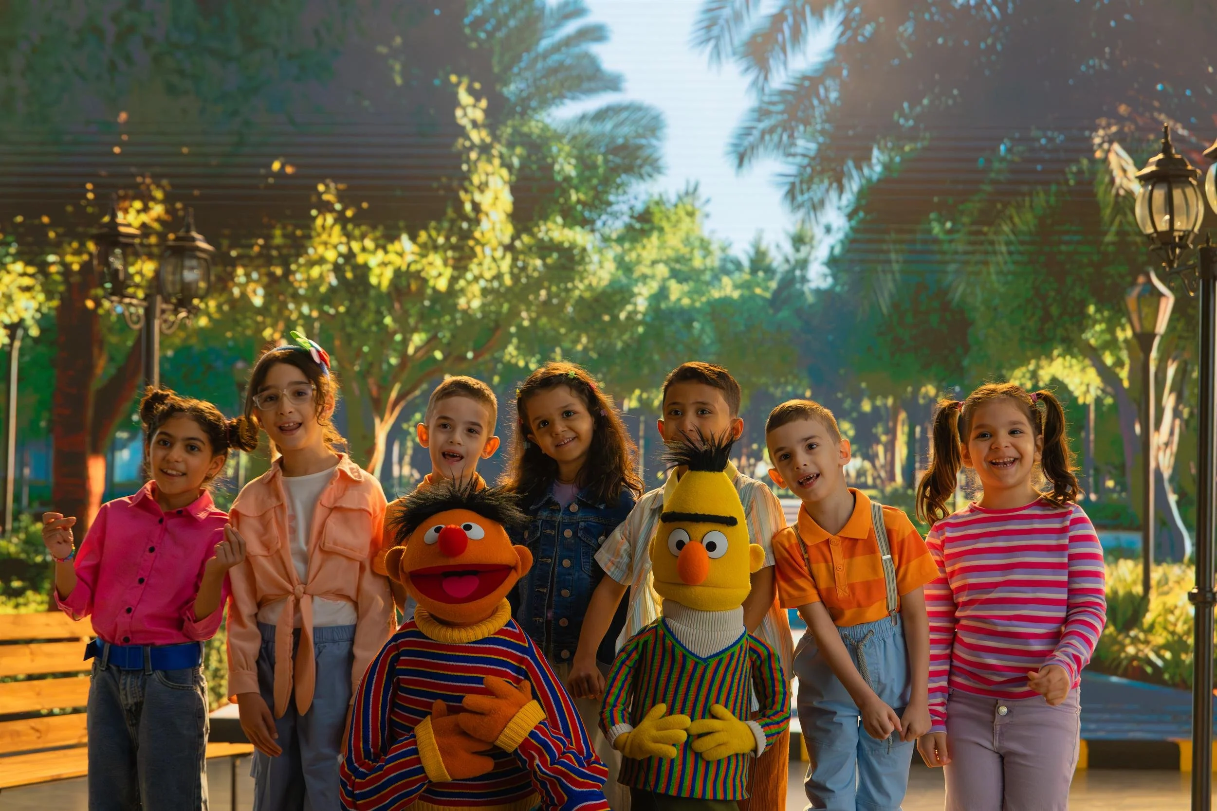 Ahlan Simsim unit still: Ernie, Bert, and a group of smiling children stand together in a sunny park setting.