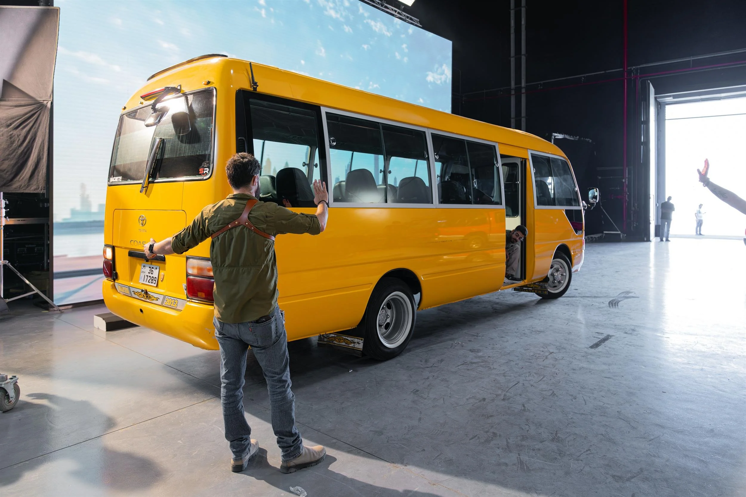 Ahlan Simsim unit still: a crew member stands behind a yellow bus on a studio floor with an LED skyline backdrop.