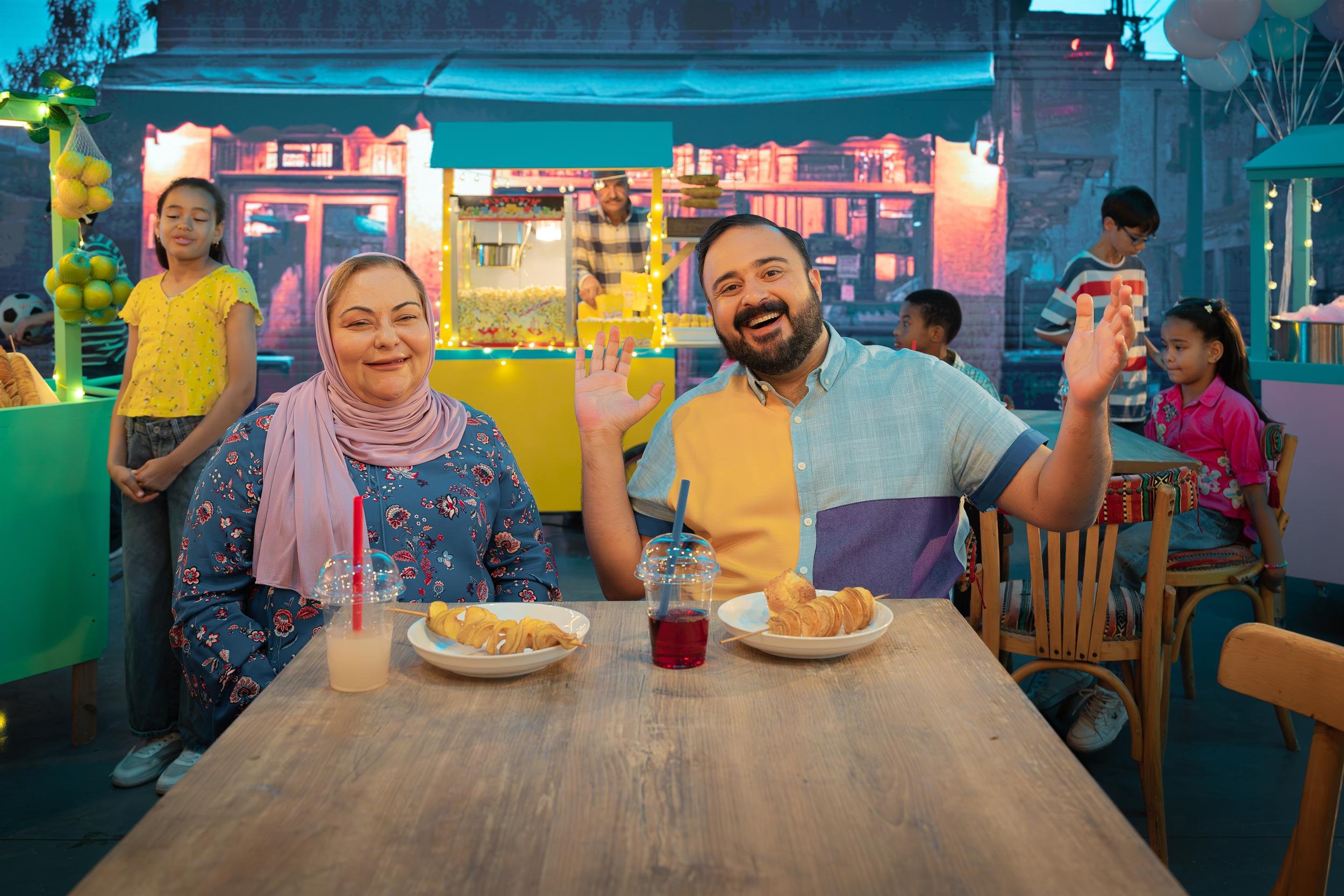 Ahlan Simsim key art: A man and woman sit at a table with snacks in a vibrant outdoor marketplace with a popcorn cart.