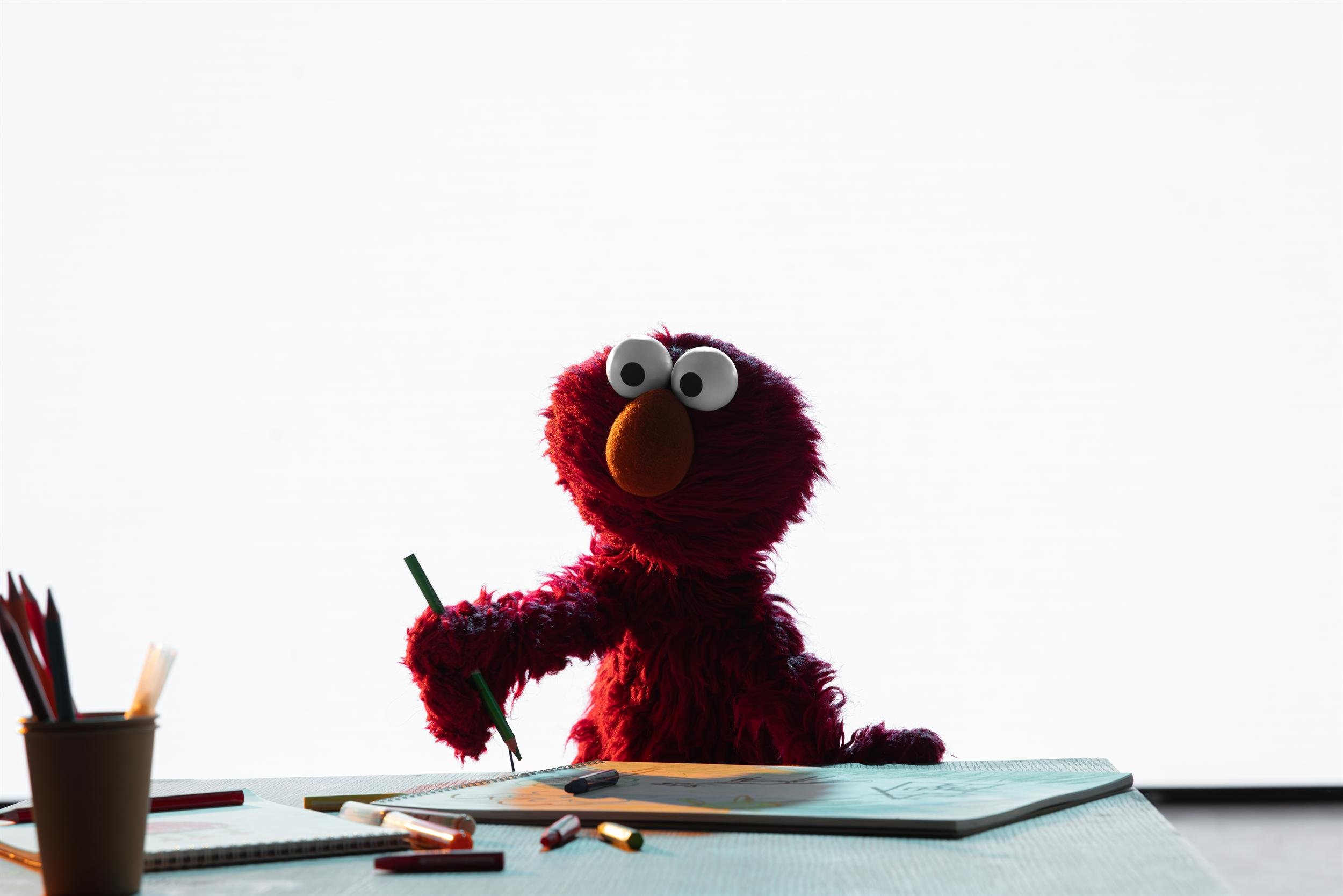 Ahlan Simsim key art: Elmo sits at a desk with colored pencils, holding a green pencil over a notebook.