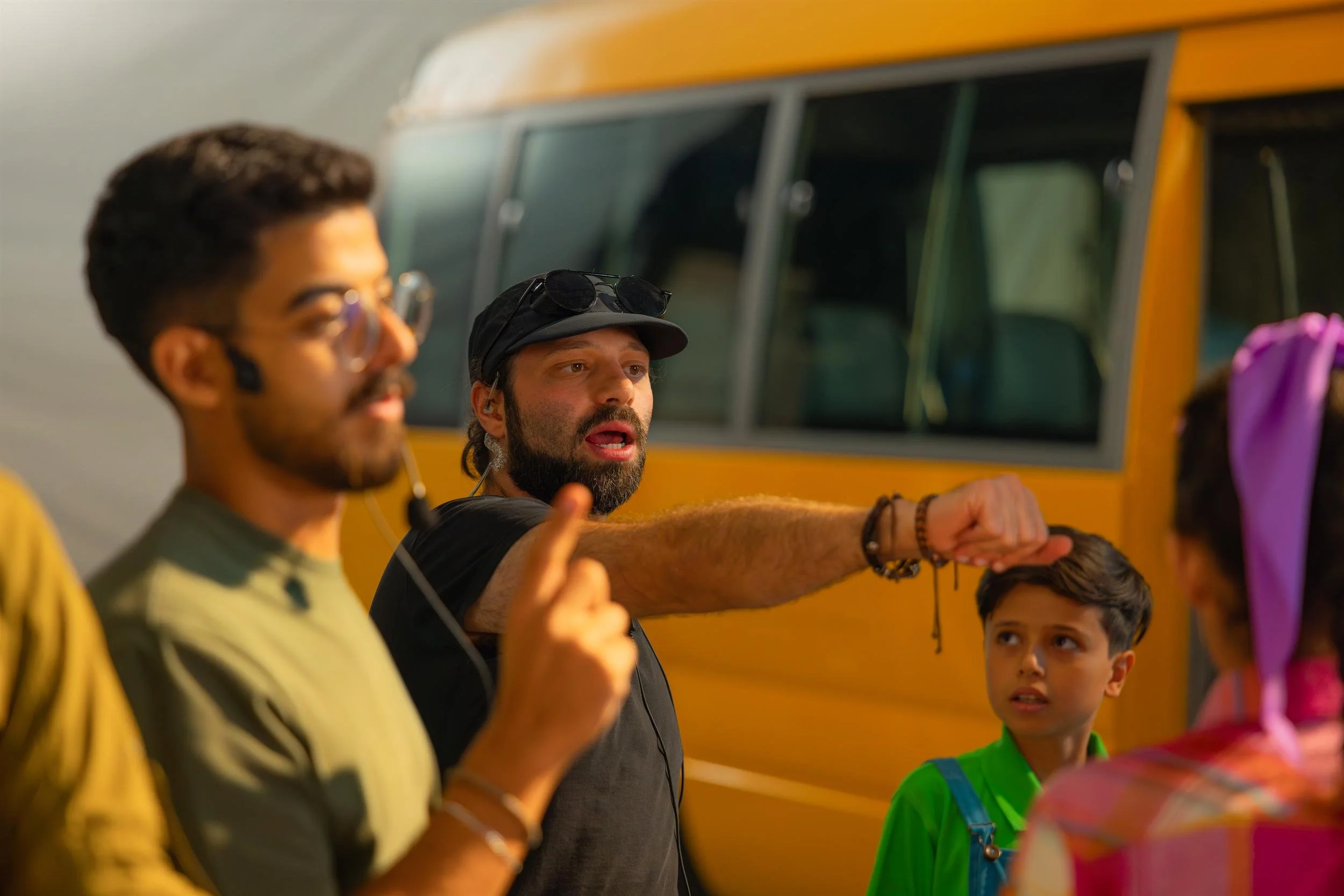 Ahlan Simsim unit still: a bearded crew member in a headset points while a boy in overalls looks on near a yellow bus.