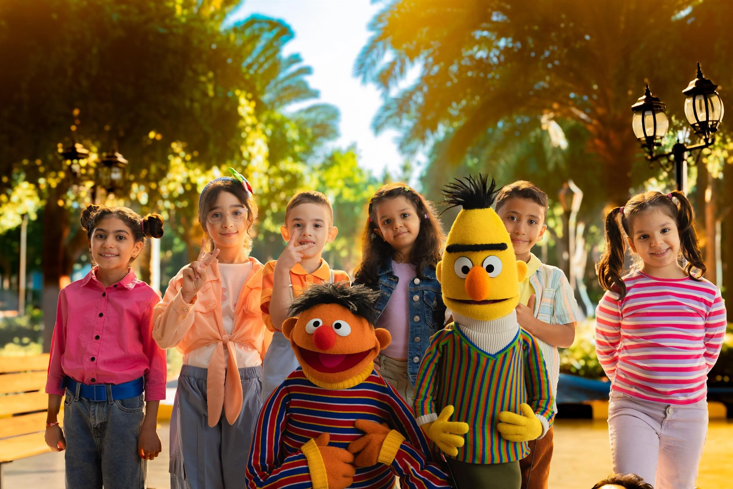 Ahlan Simsim unit still: Ernie, Bert, and six children pose and smile in a sun-drenched park.