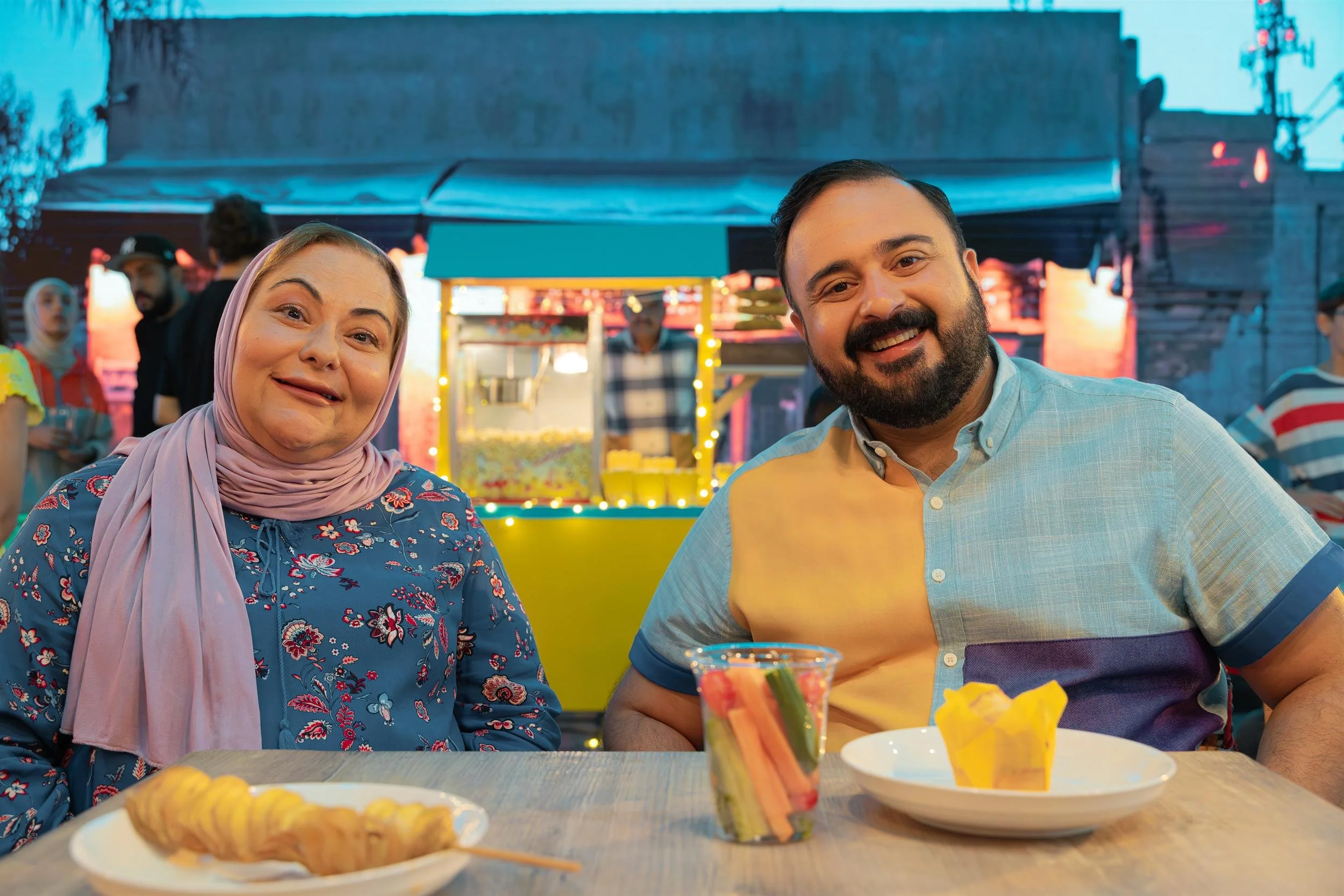 Ahlan Simsim unit still: a man and a woman smile at a table with snacks in front of a popcorn stand.