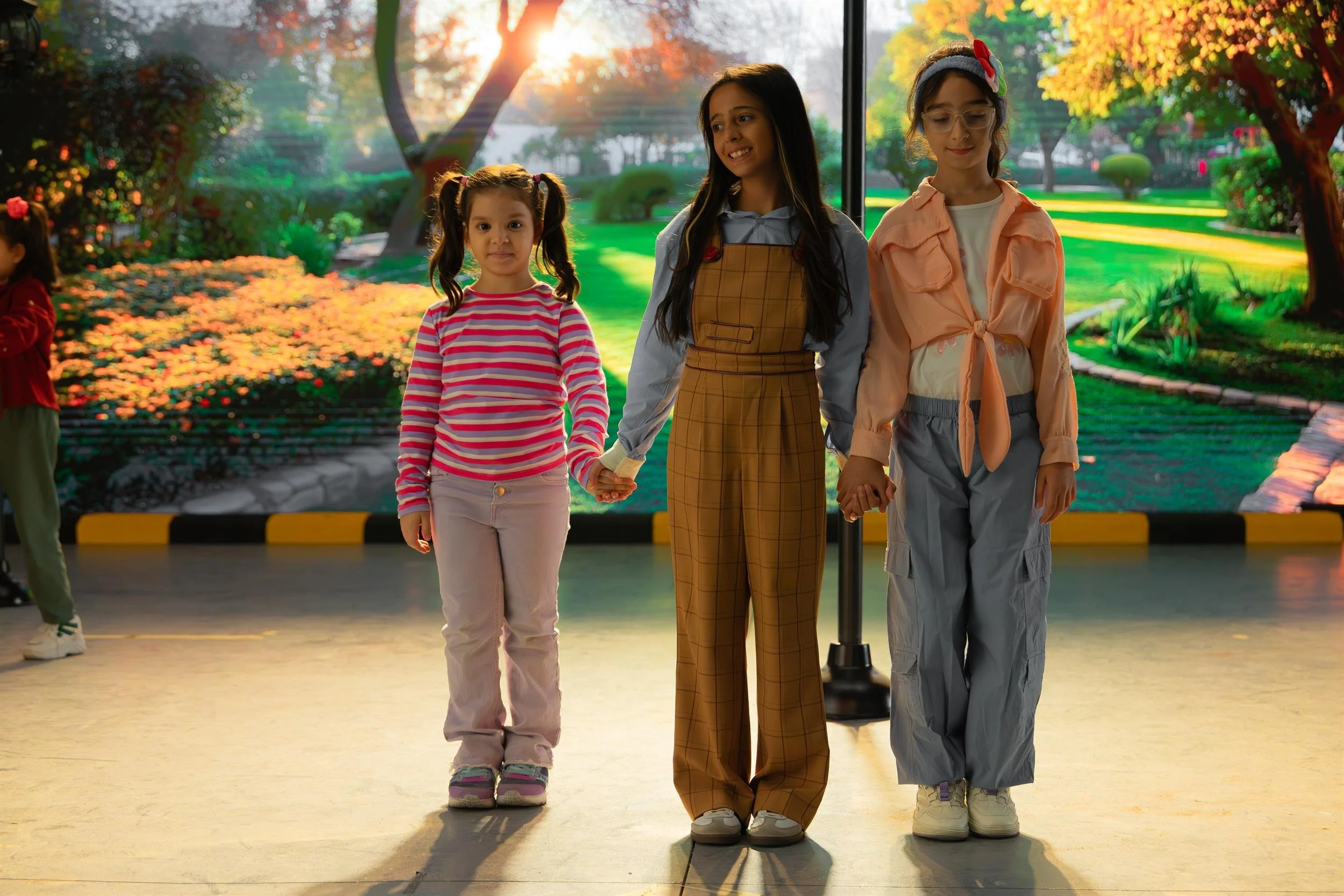 Ahlan Simsim unit still: three girls hold hands on a grey studio floor before a sunlit park backdrop.