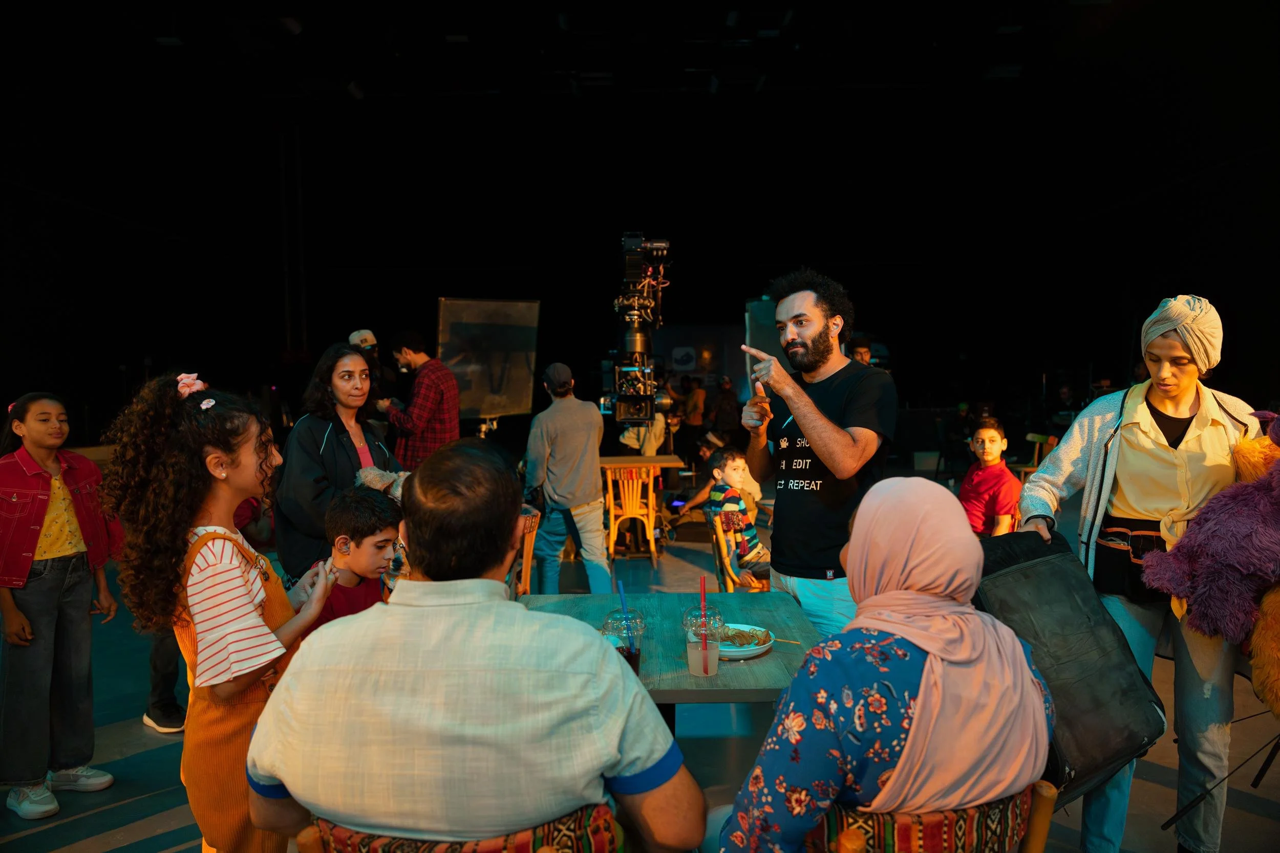 Ahlan Simsim unit still: a crew member directs people at a table on a dark set with a purple Muppet nearby.