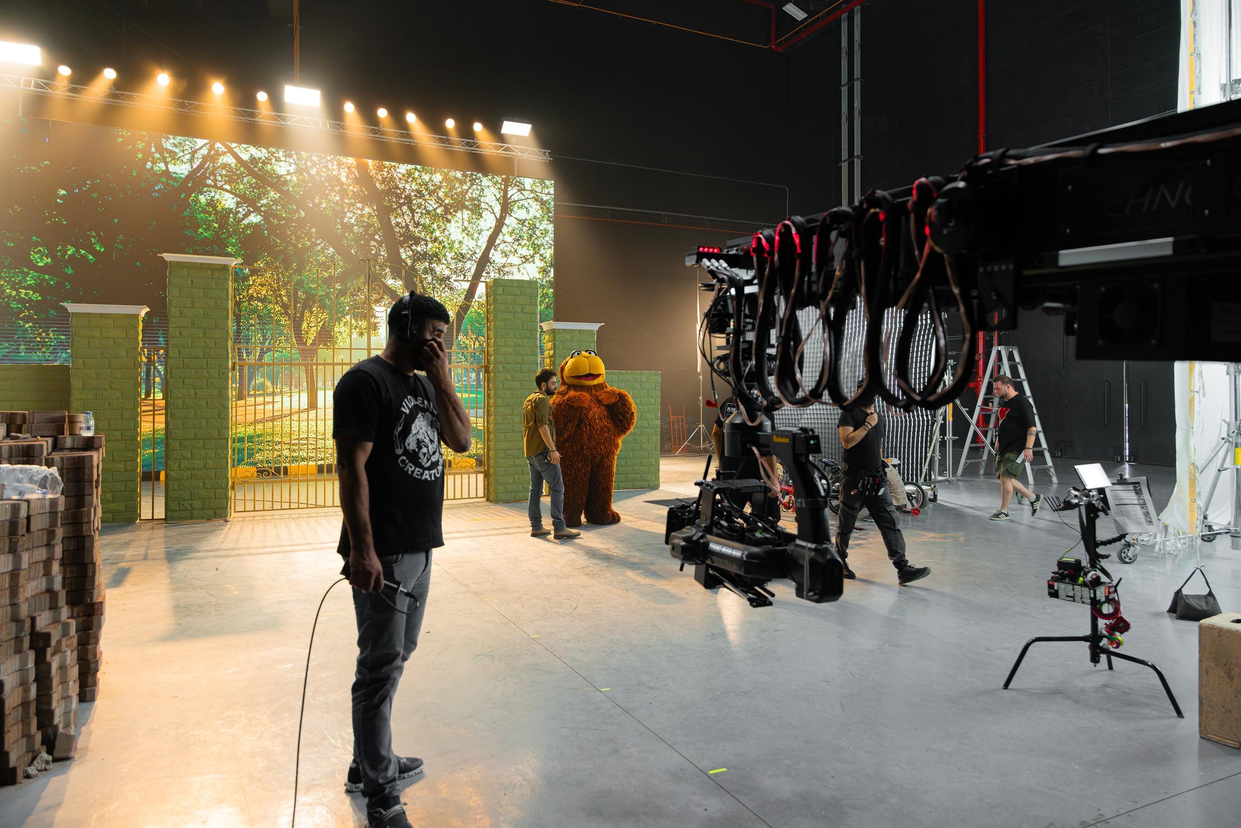Ahlan Simsim unit still: A brown Muppet stands on a park-gate set while a camera crane and crew fill the studio.