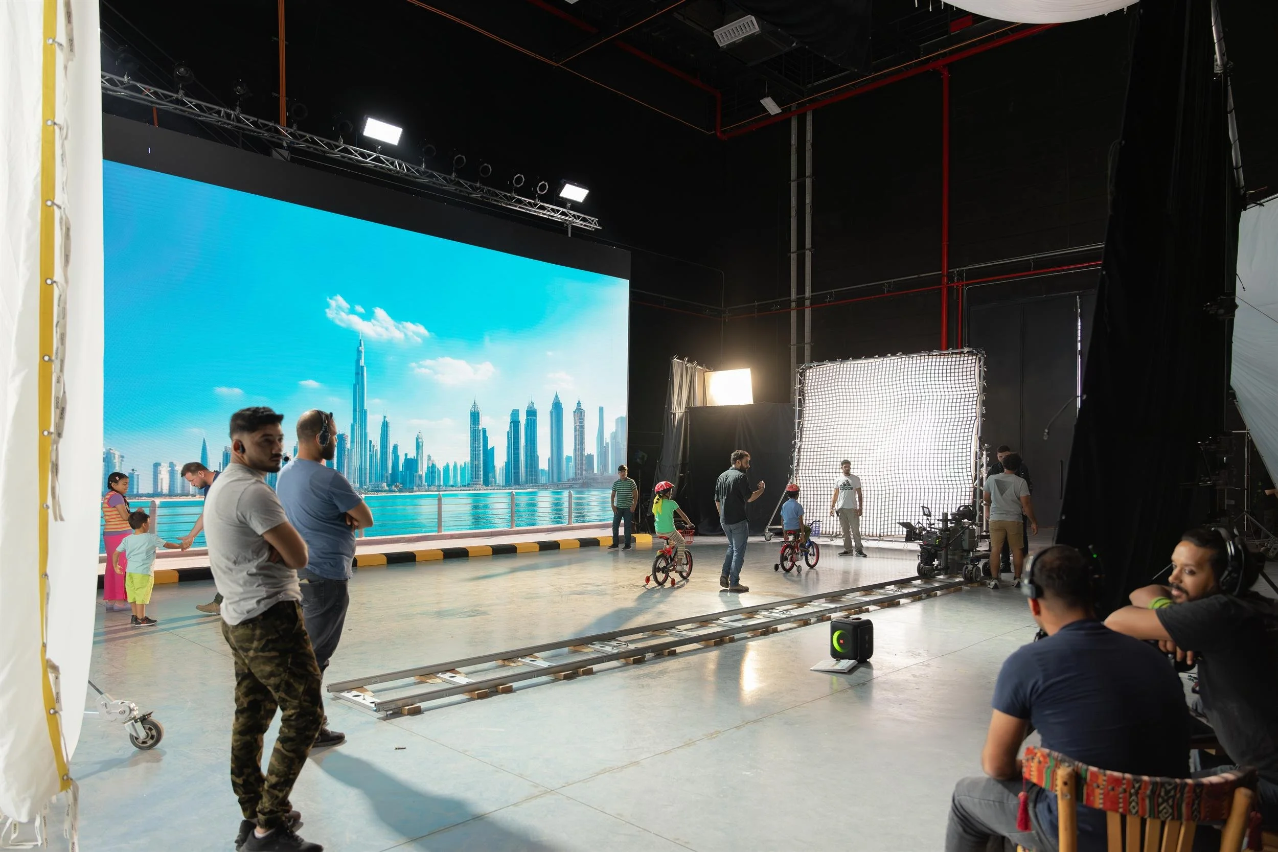 Ahlan Simsim unit still: crew film children on bicycles in a studio with an LED city skyline.