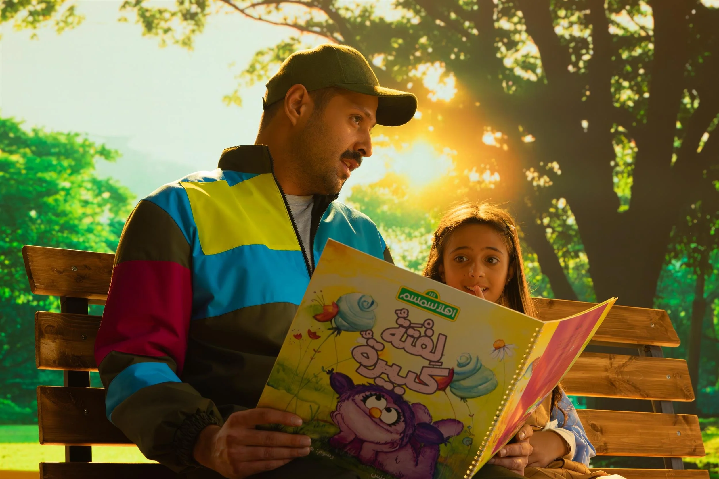 Ahlan Simsim key art: a man in a cap reading a picture book to a young girl on a park bench