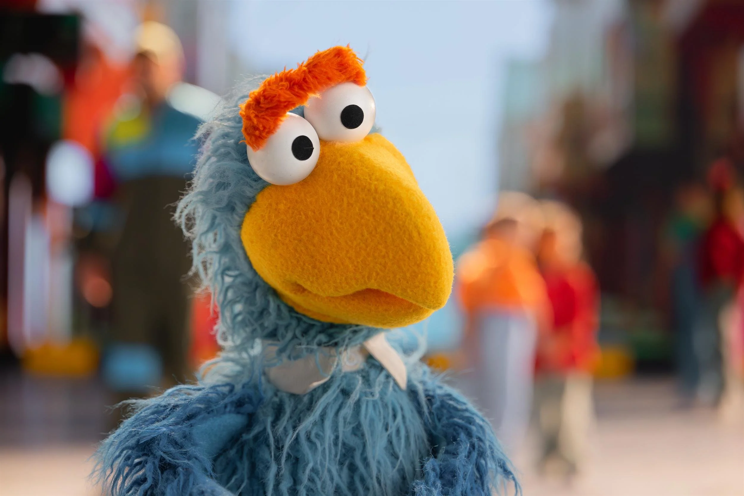 Close-up of a blue feathered Muppet with orange hair on a busy street set
