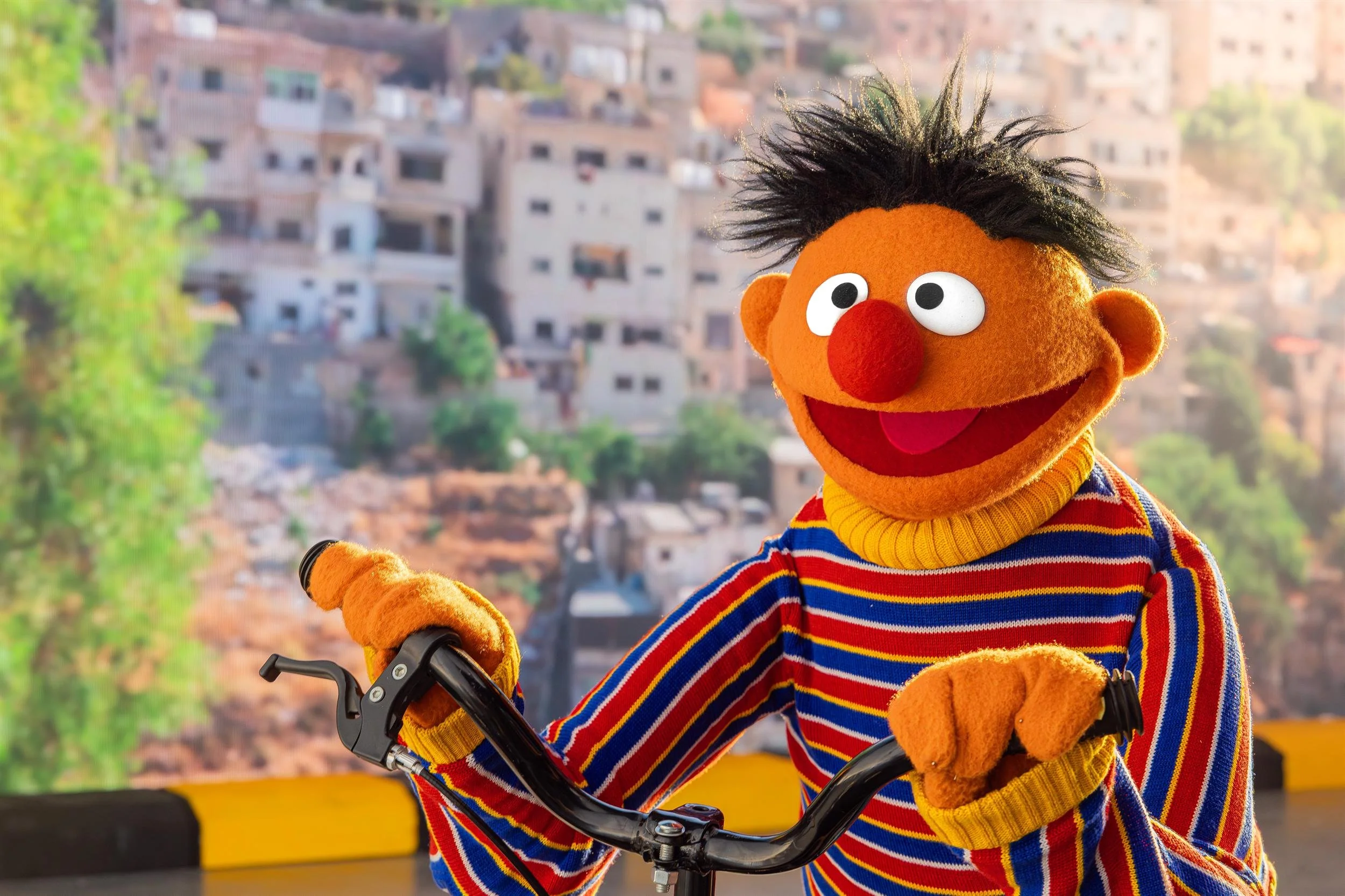 Ernie Muppet riding a bicycle with a Jordanian hillside town behind him