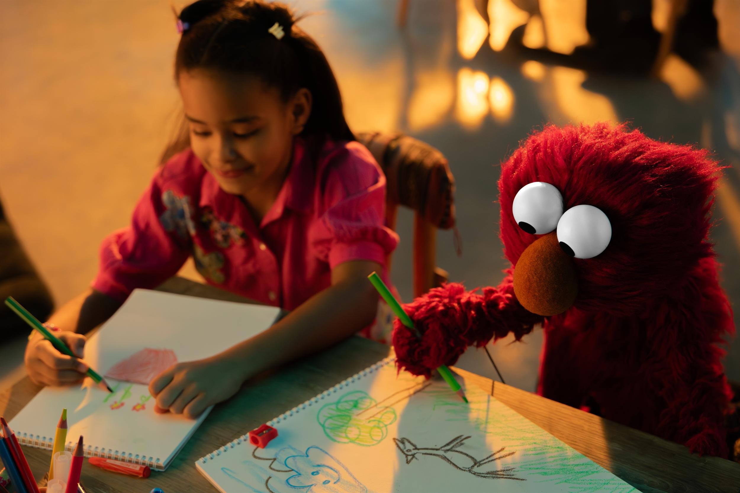 Young girl drawing at a table beside Elmo in warm lamplight