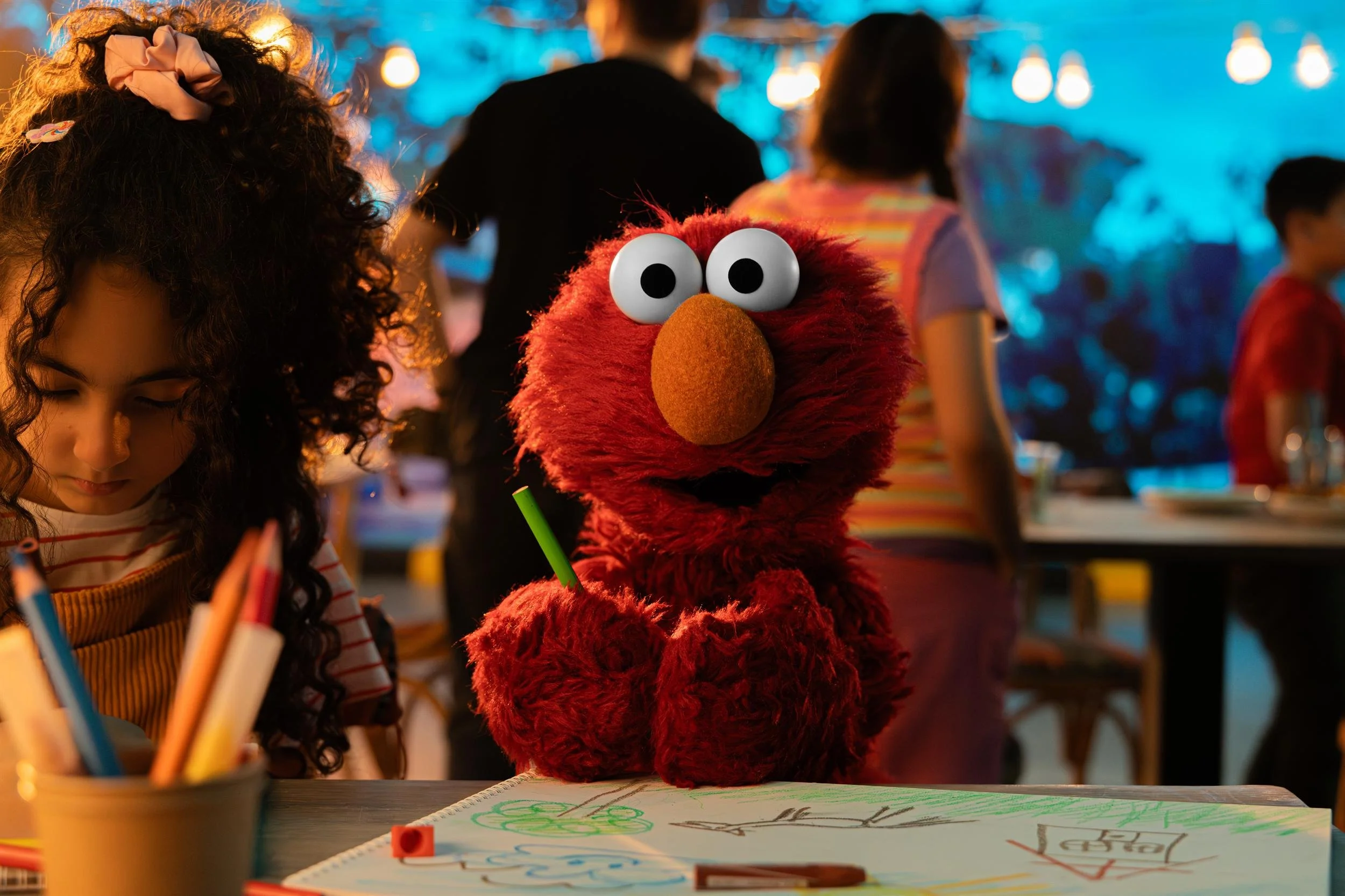 Ahlan Simsim key art: Elmo sitting at a classroom table beside a girl drawing with coloured pencils