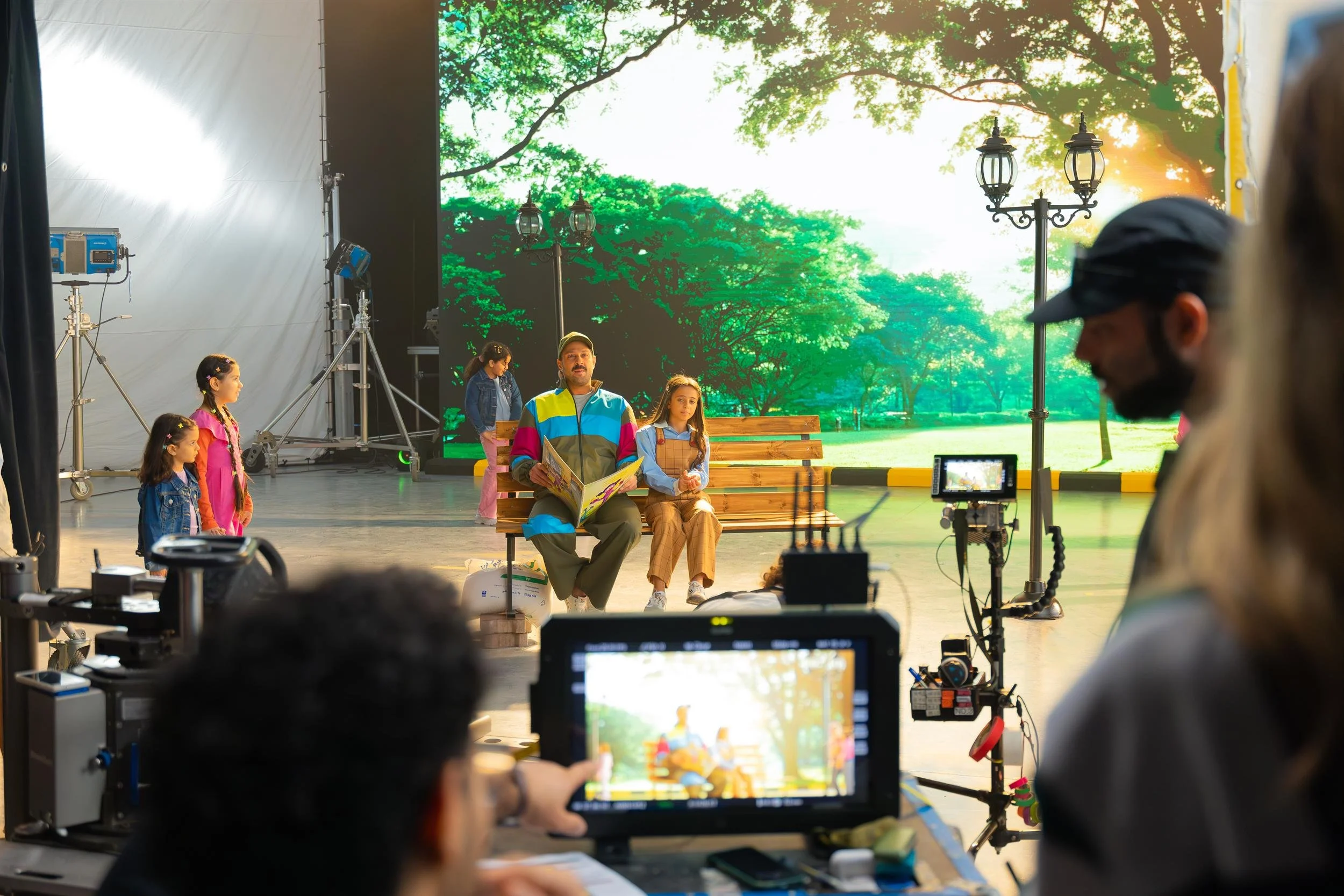 Ahlan Simsim unit still: Crew film a man and children on a park-bench set in front of a large LED backdrop.