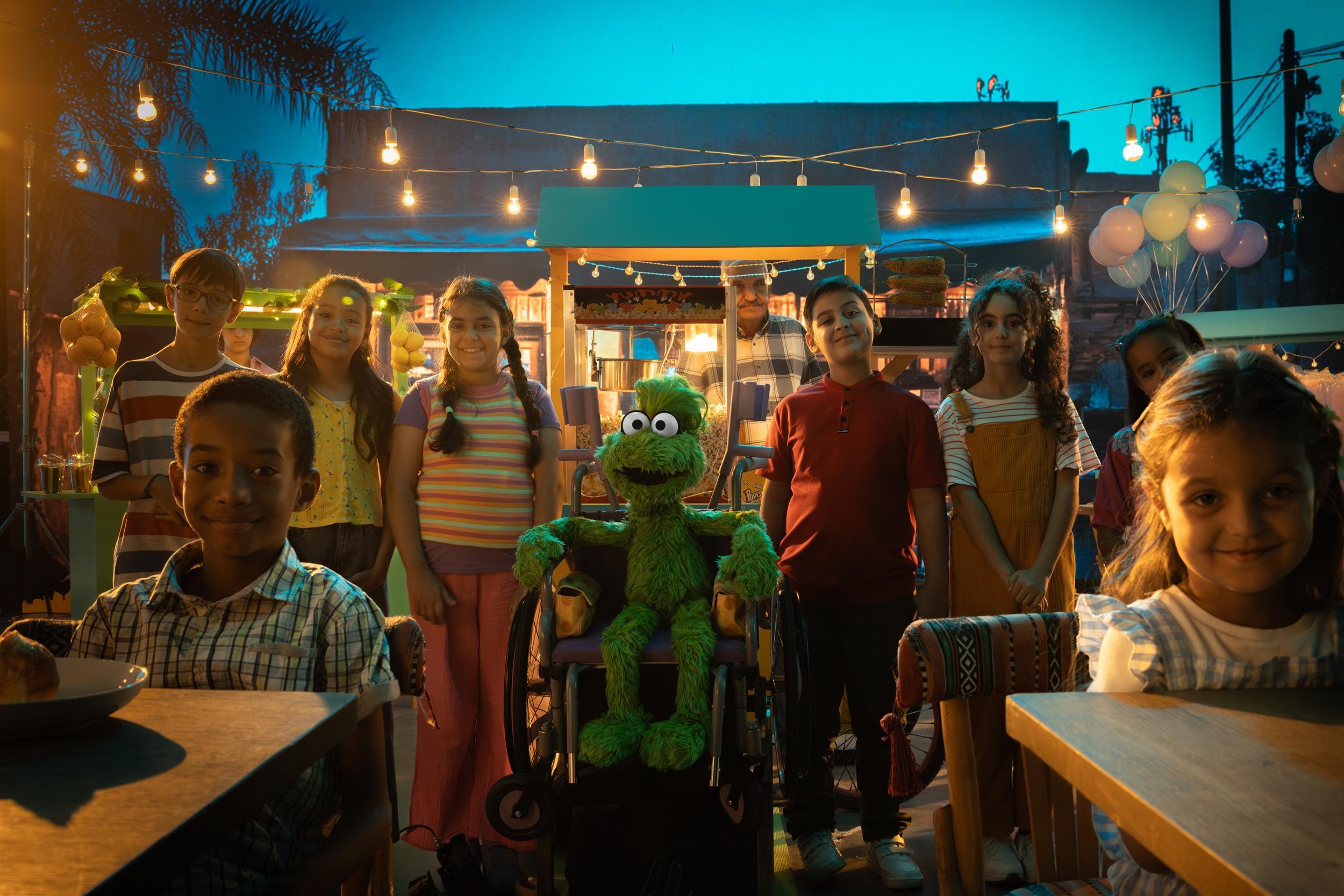 Ahlan Simsim key art: Children and a green Muppet in a wheelchair pose in an outdoor night market under string lights.
