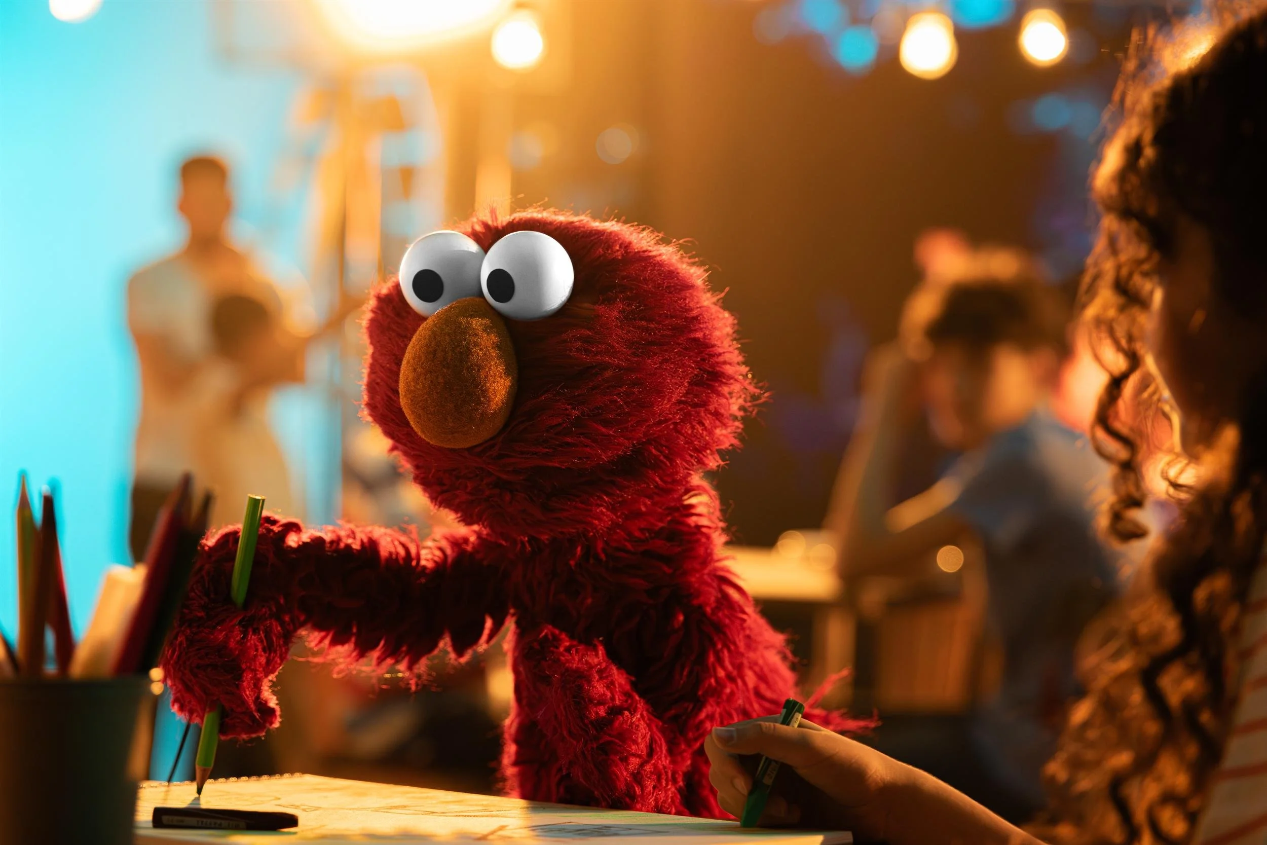 Ahlan Simsim key art: Elmo draws with a child using colored pencils at a table in a warm indoor set.