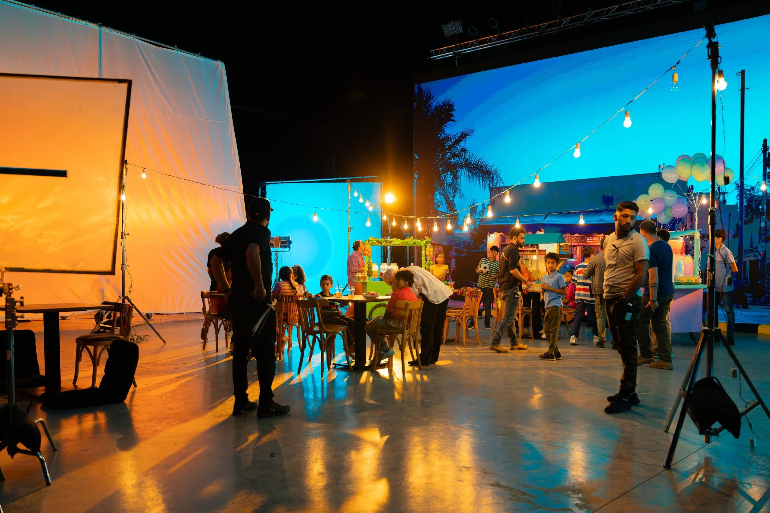 Ahlan Simsim unit still: a wide studio view of a night-market set, LED street backdrop, and children at tables.