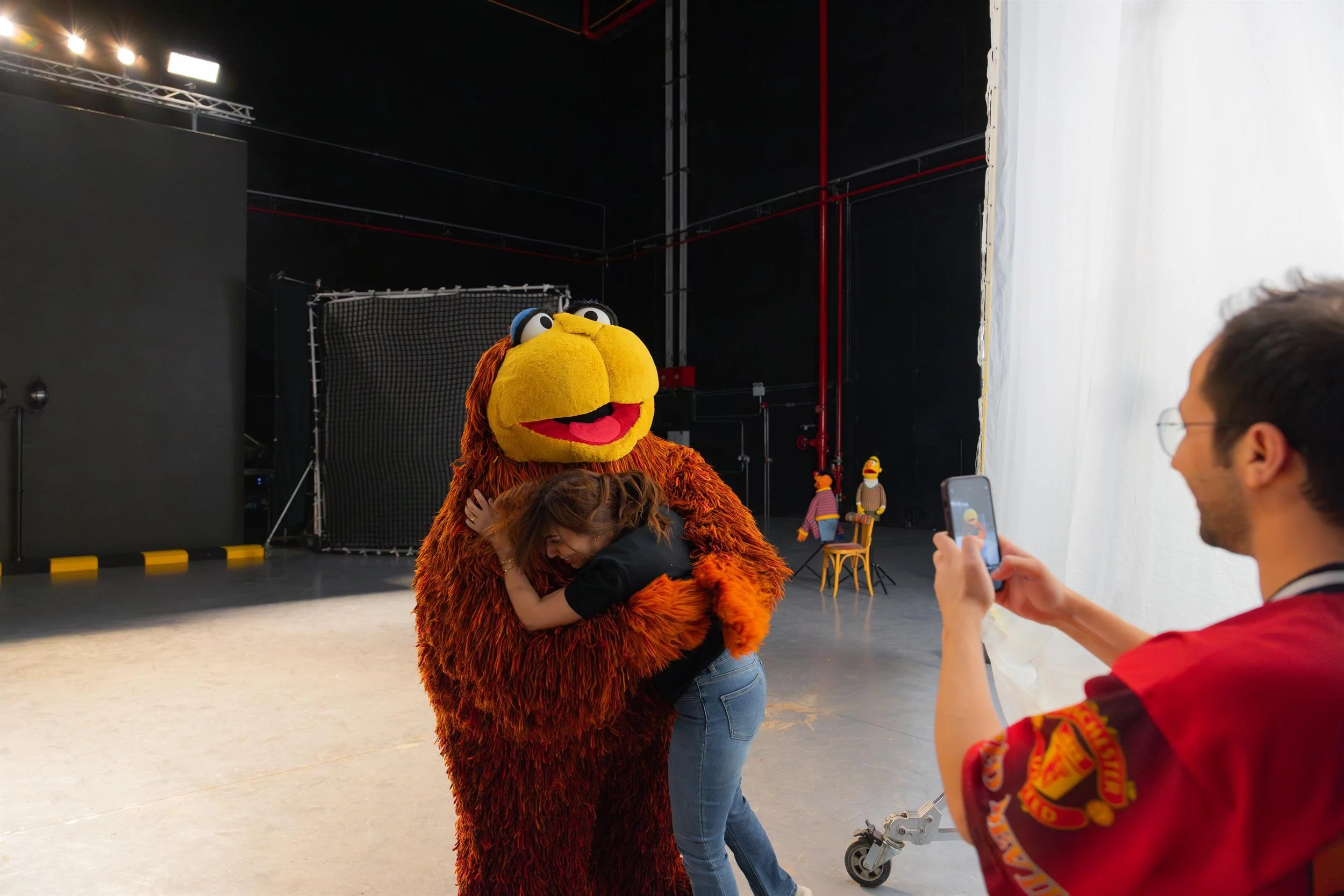 Ahlan Simsim unit still: a woman hugs a large brown Muppet while a man photographs them; Ernie and Bert sit in the background.