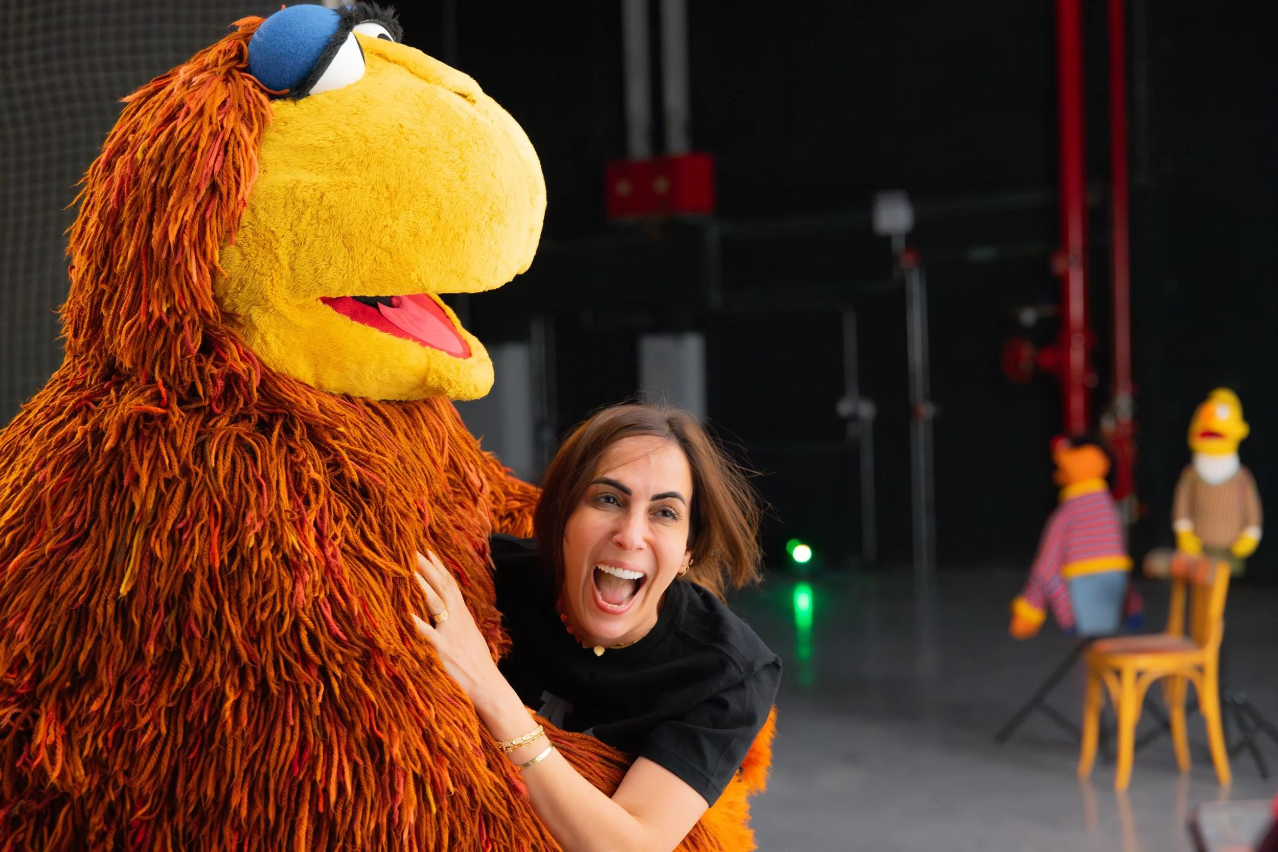 Ahlan Simsim unit still: a smiling woman hugs a large brown Muppet in a dark studio with Ernie and Bert behind them.