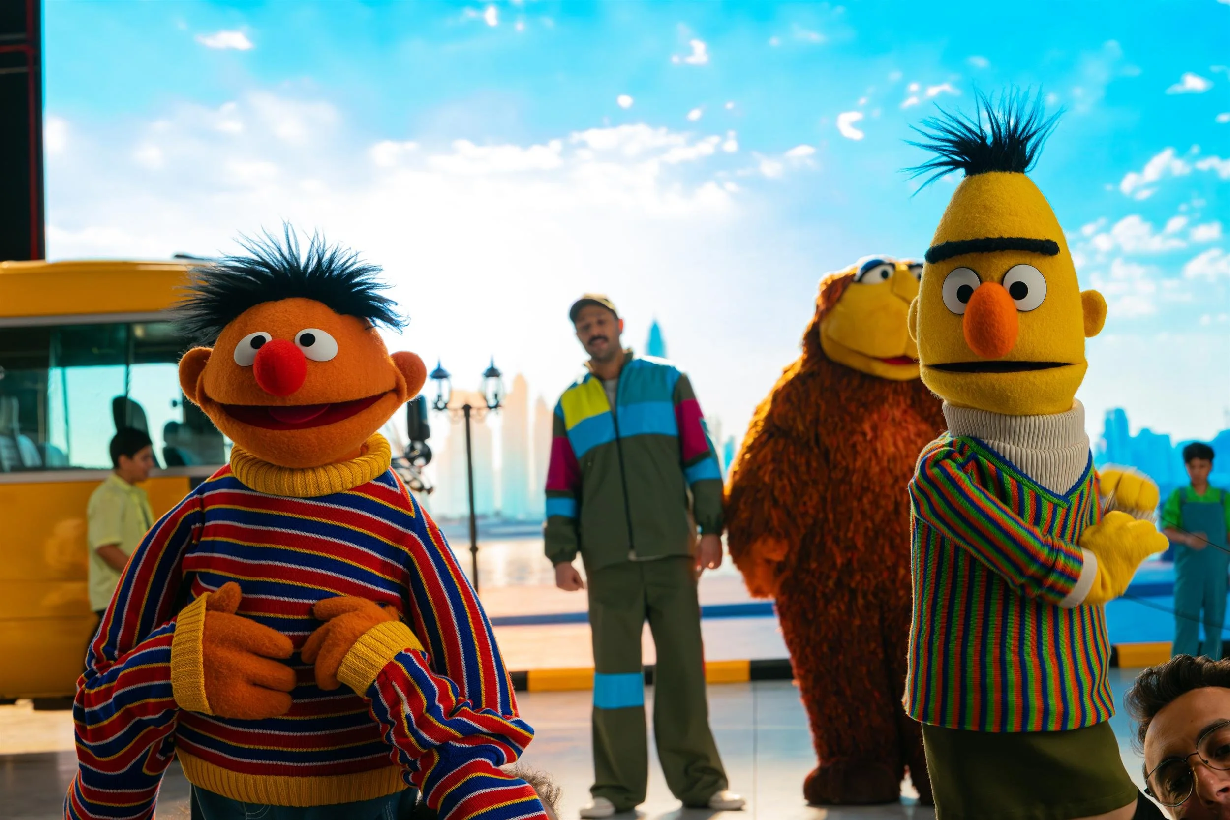 Ahlan Simsim unit still: Ernie, Bert, and a large brown Muppet stand by a yellow bus against an LED city skyline.