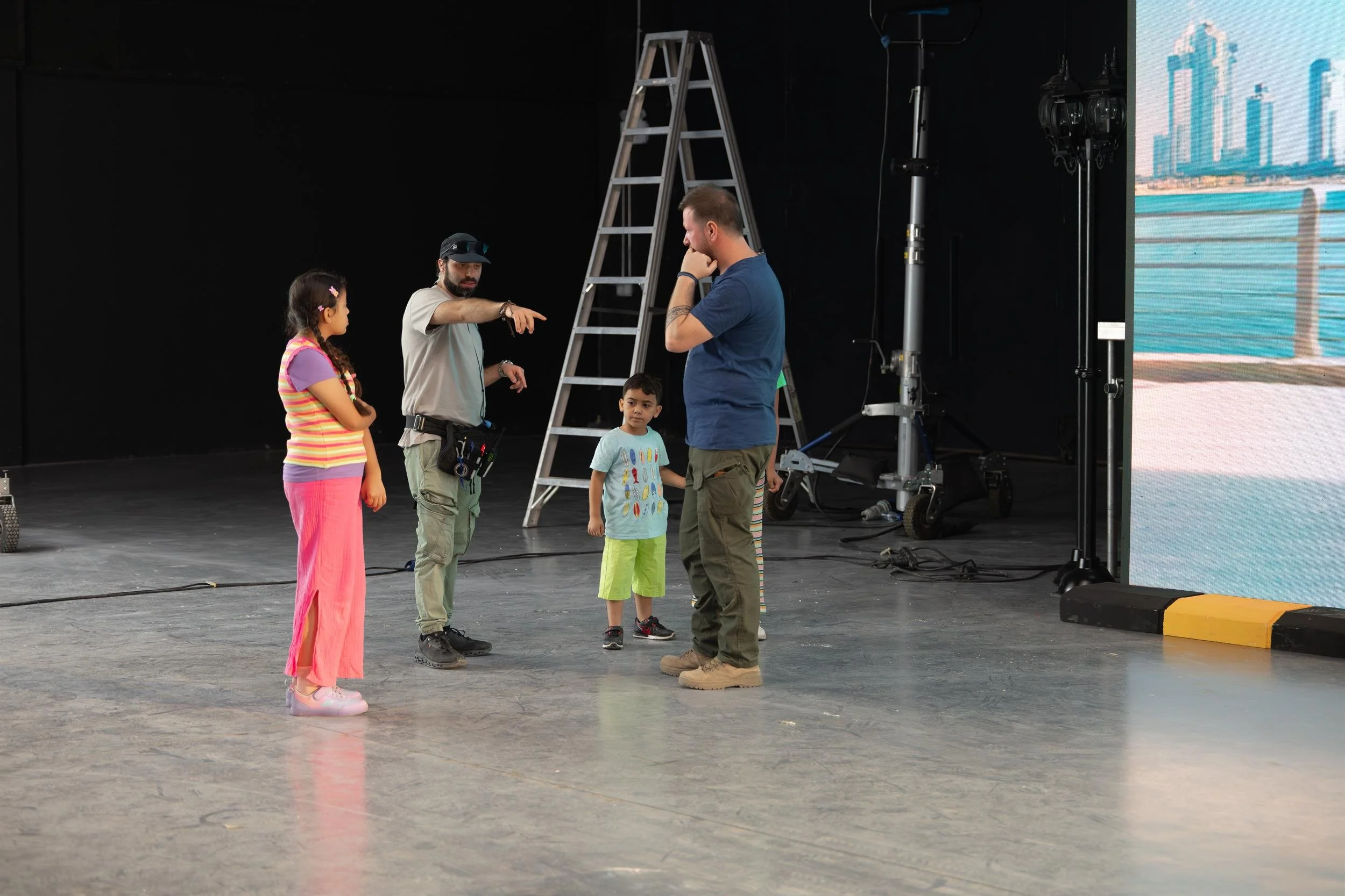 Ahlan Simsim unit still: crew members direct two children on a studio set before an LED waterfront skyline.Ahlan Simsim unit still: crew members direct two children on a studio set before an LED waterfront skyline.