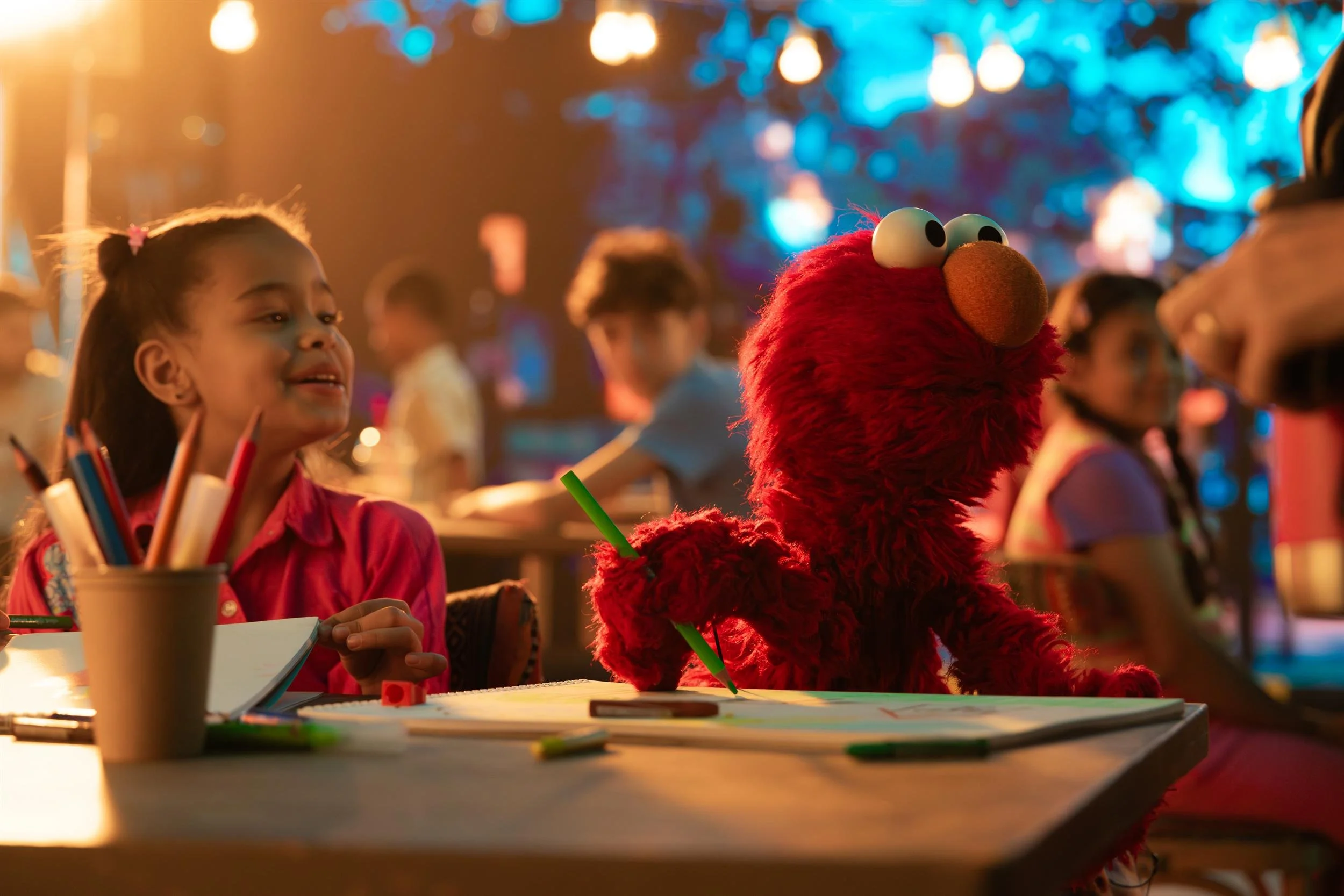 Ahlan Simsim unit still: Elmo and a smiling girl sit at a table drawing with markers and colored pencils.
