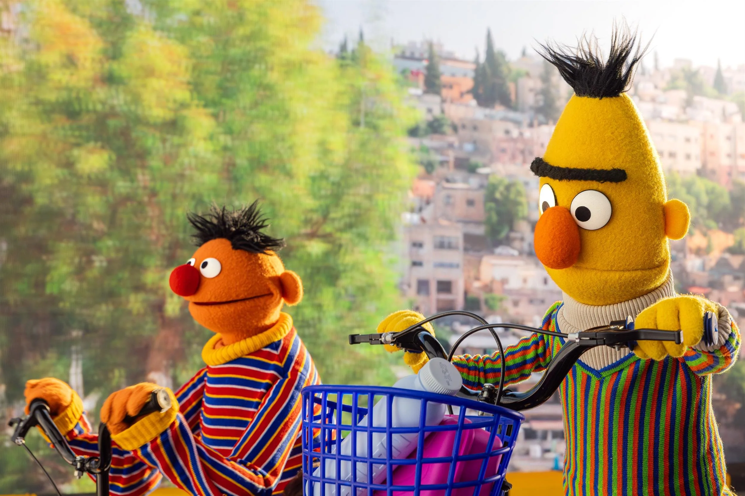 Ahlan Simsim key art: Bert and Ernie ride bicycles in front of a sunlit hillside city.