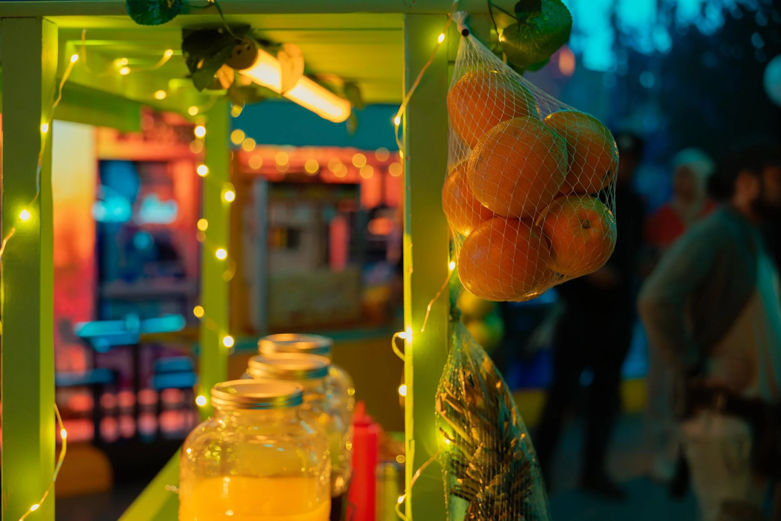 Ahlan Simsim unit still: hanging oranges and juice jars on a green night-market stall wrapped in string lights.