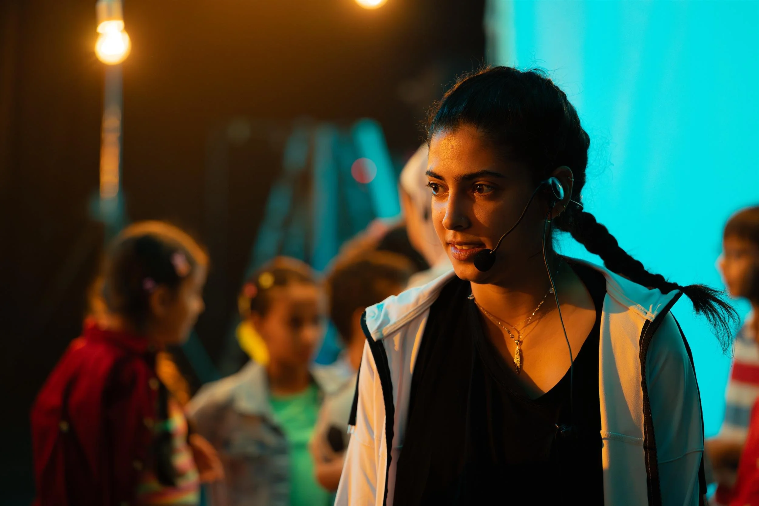 Ahlan Simsim unit still: a crew member in a headset stands on set with children blurred behind her.