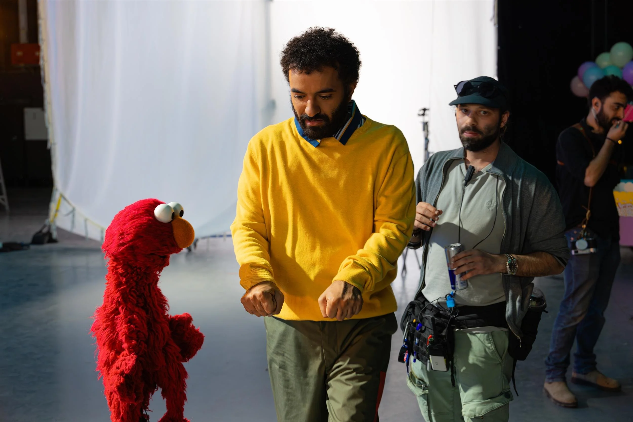 Ahlan Simsim unit still: Elmo looks up at a man in a yellow sweater while a crew member stands nearby.
