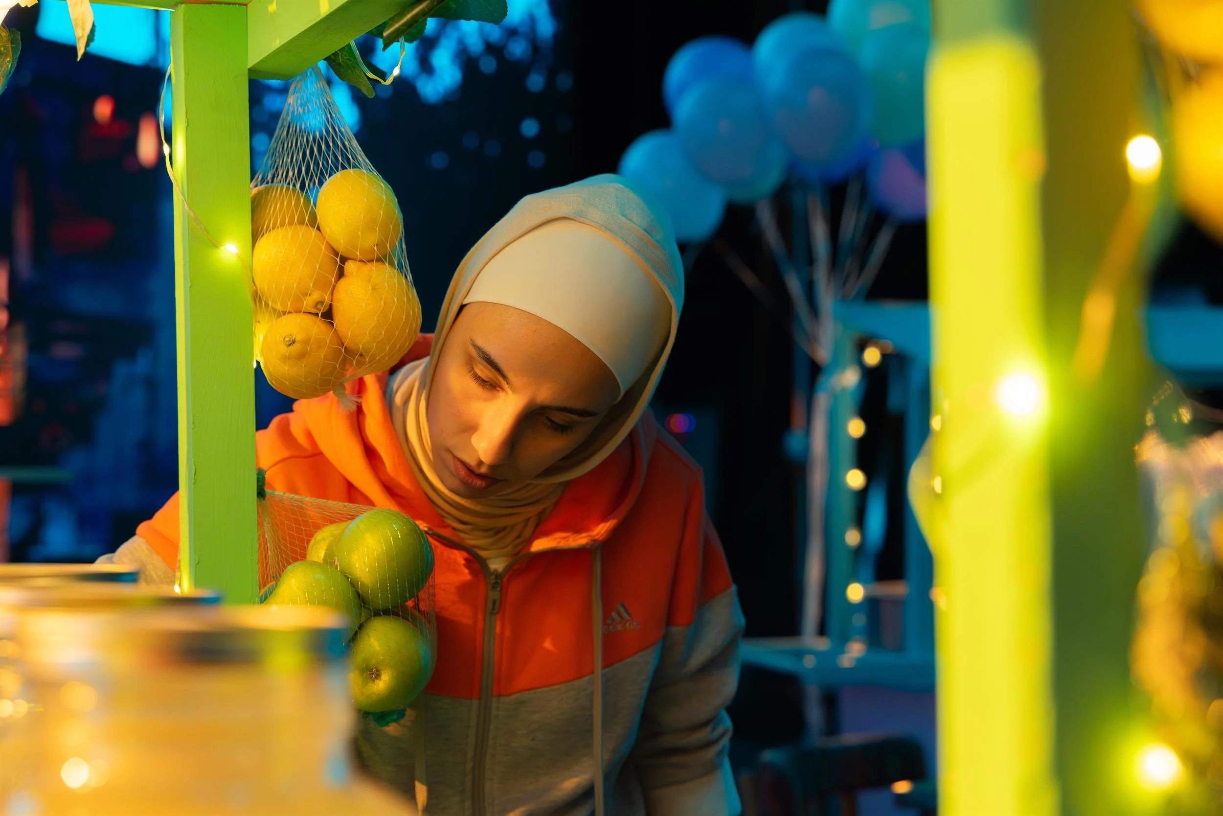 Ahlan Simsim unit still: a woman in a hijab looks down at a green market stall hung with lemons and apples.