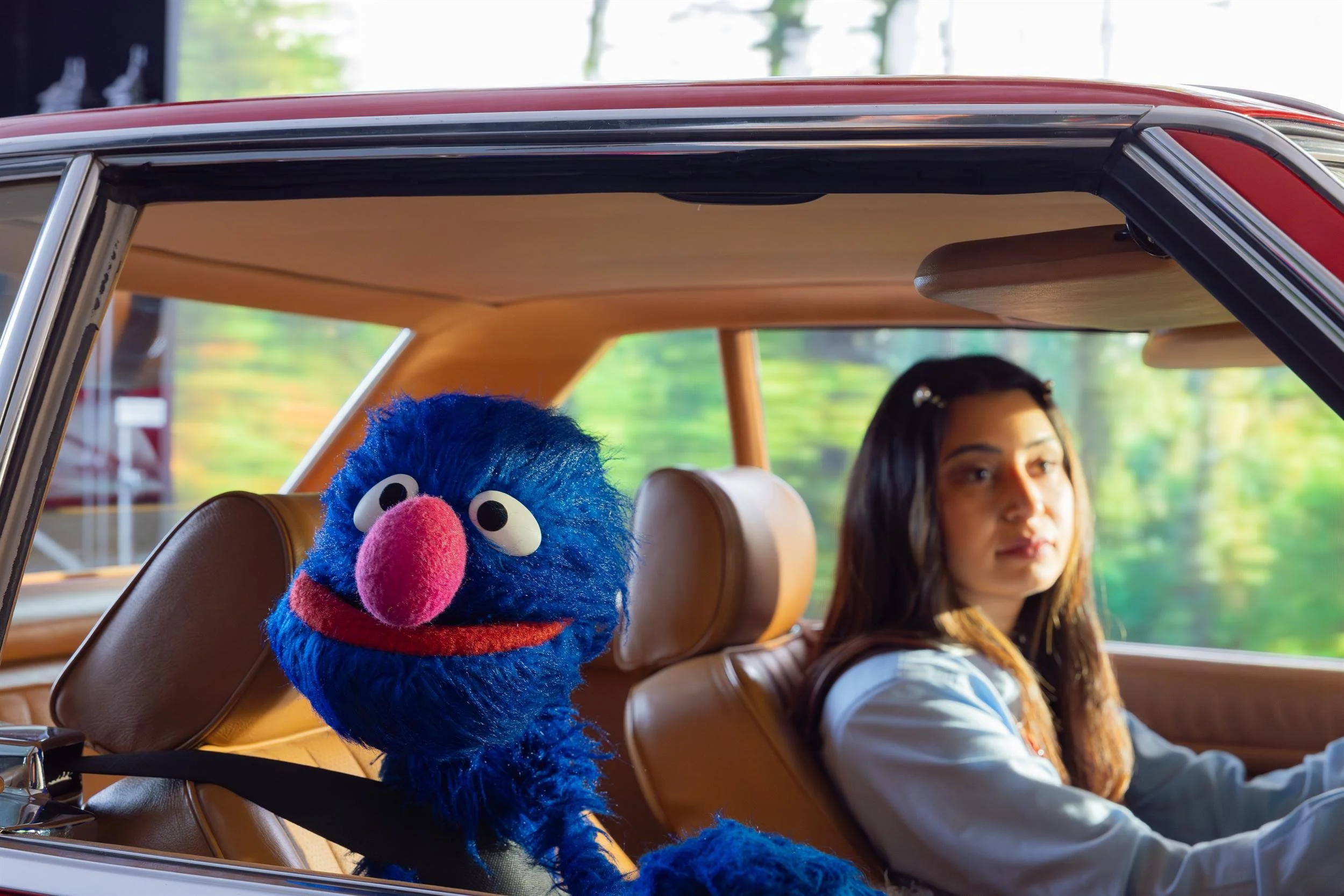 Ahlan Simsim unit still: a blue Muppet buckled into the passenger seat of a vintage red car next to a young woman.