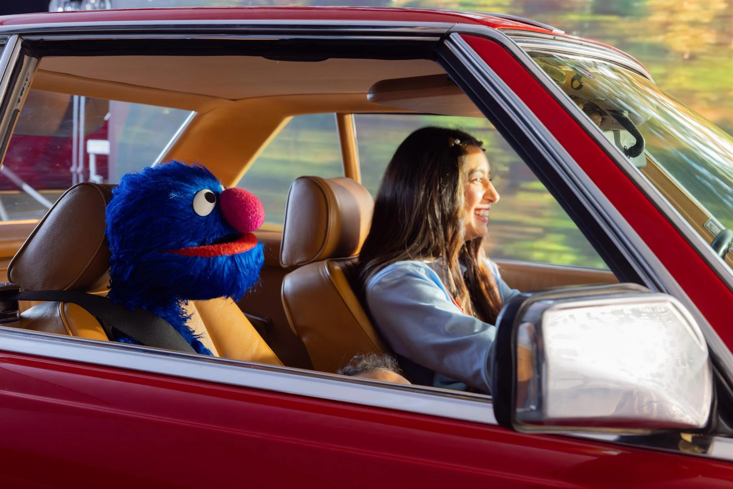 Ahlan Simsim unit still: a smiling woman drives a vintage red car with a blue Muppet buckled in the back seat.