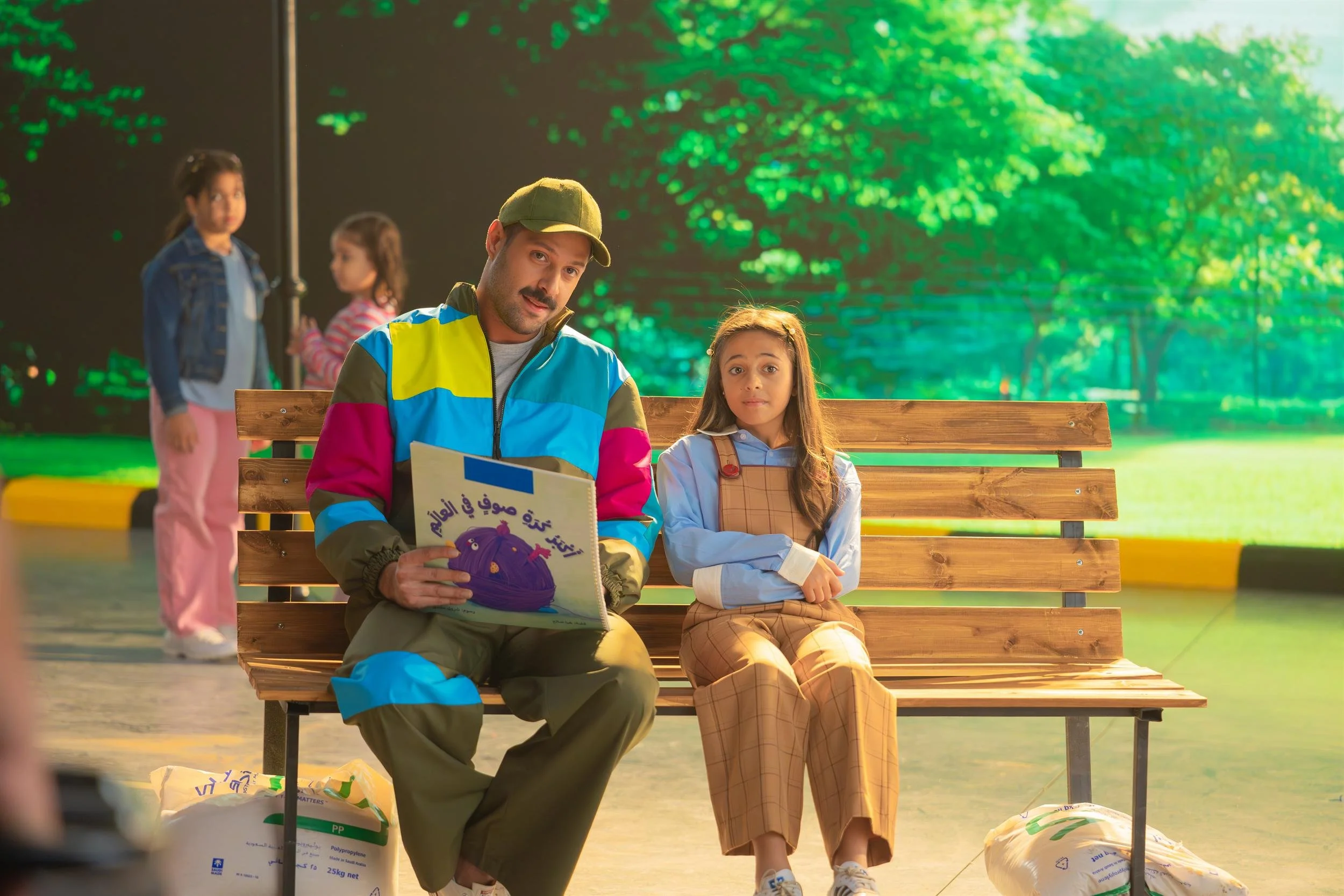 Ahlan Simsim unit still: A man in a colorful jacket and a young girl in overalls sit together on a wooden park bench.