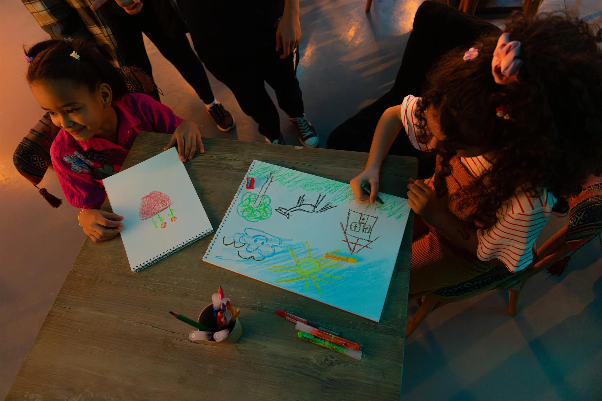 Ahlan Simsim unit still: two girls sit at a wooden table drawing with crayons as adults look on.