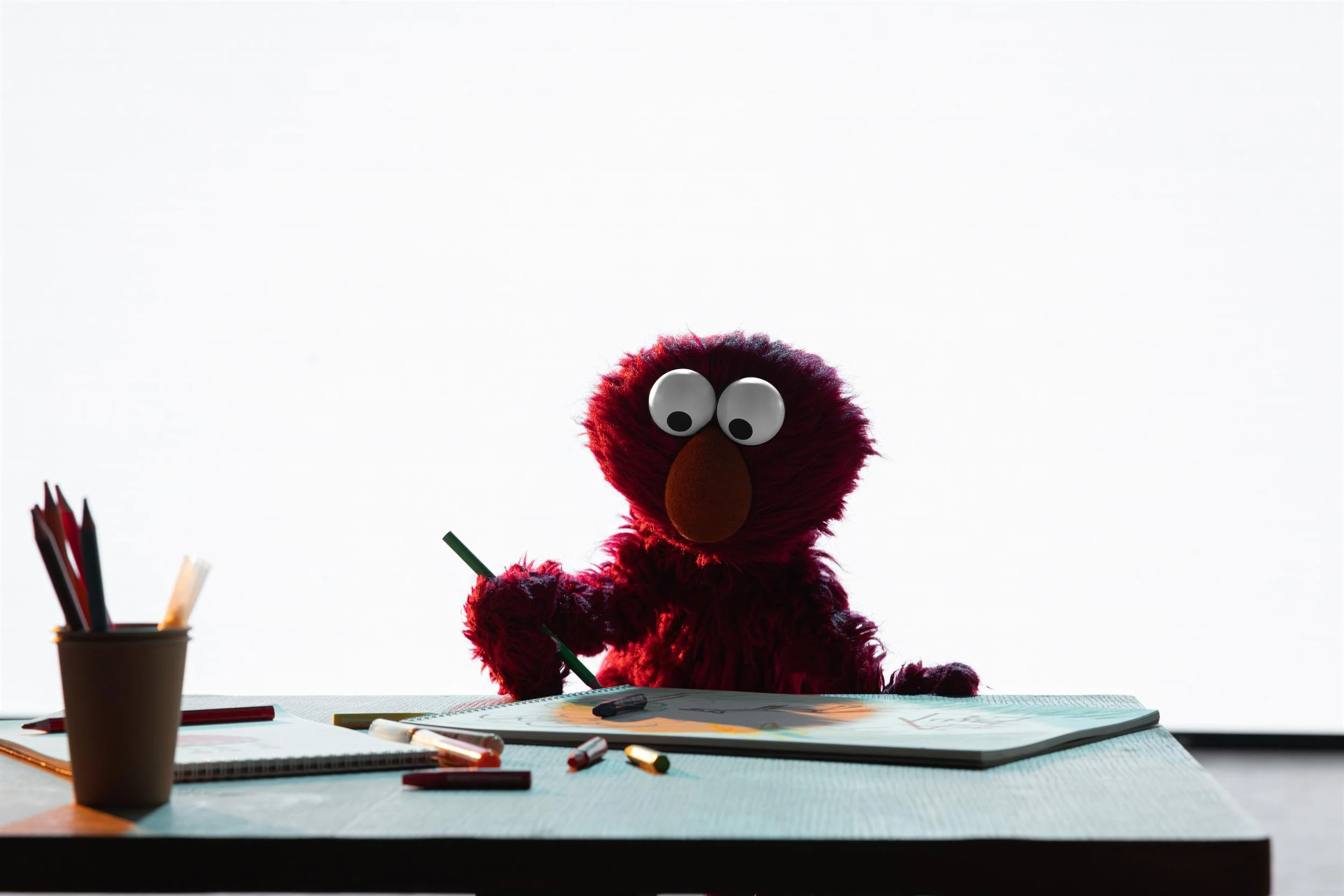 Ahlan Simsim key art: Elmo sits at a light blue desk with scattered pencils and a sketchbook against a bright studio backdrop.