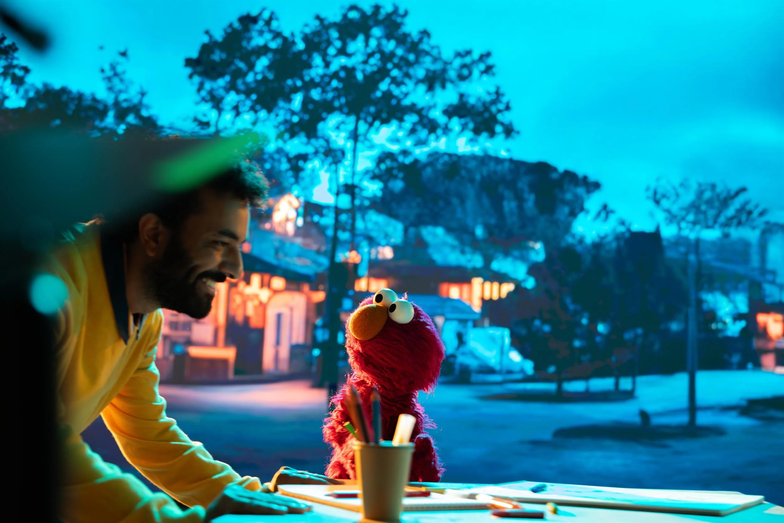 Ahlan Simsim unit still: a man in a yellow shirt smiles at Elmo at a desk with art supplies.