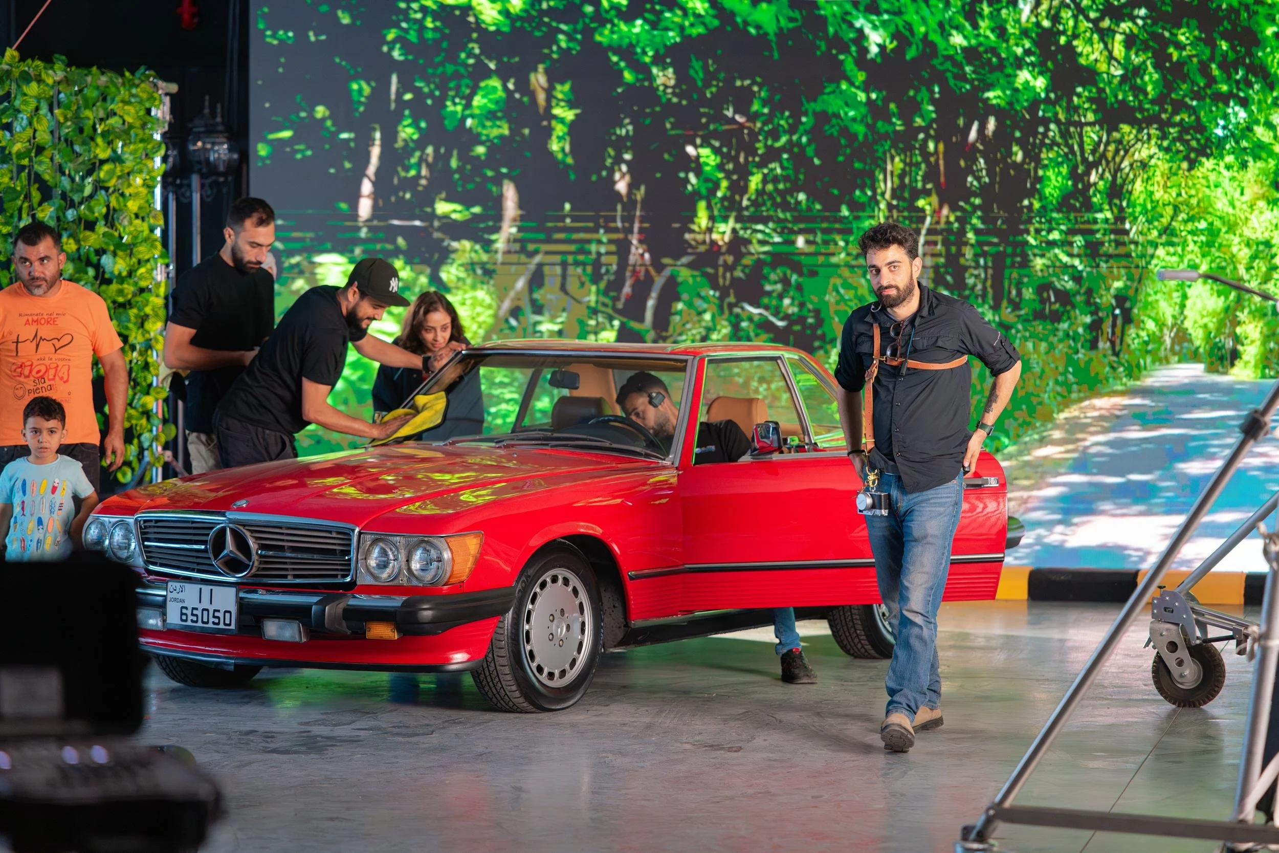 Ahlan Simsim unit still: a vintage red car sits on a studio floor before an LED forest road, with crew around it.
